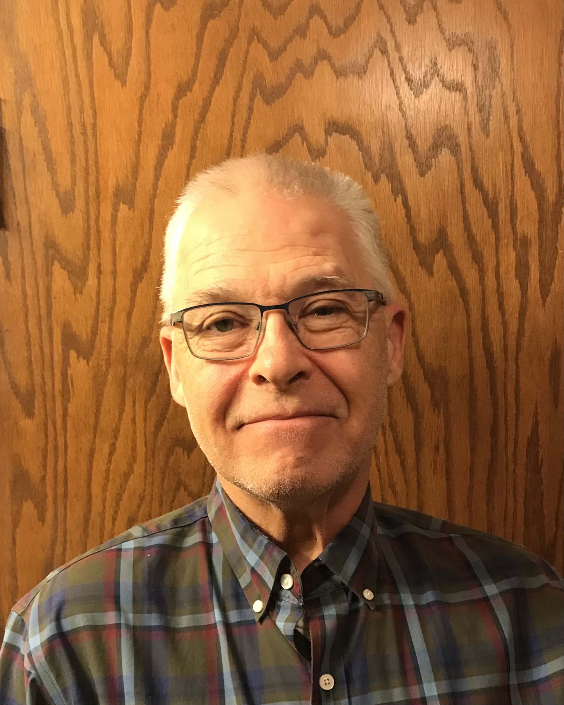 A middle-aged man with glasses and gray hair, smiling, standing in front of a wooden wall, wearing a plaid shirt.