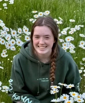 A smiling girl with long braided hair sitting outdoors in a field of white daisies and green grass.