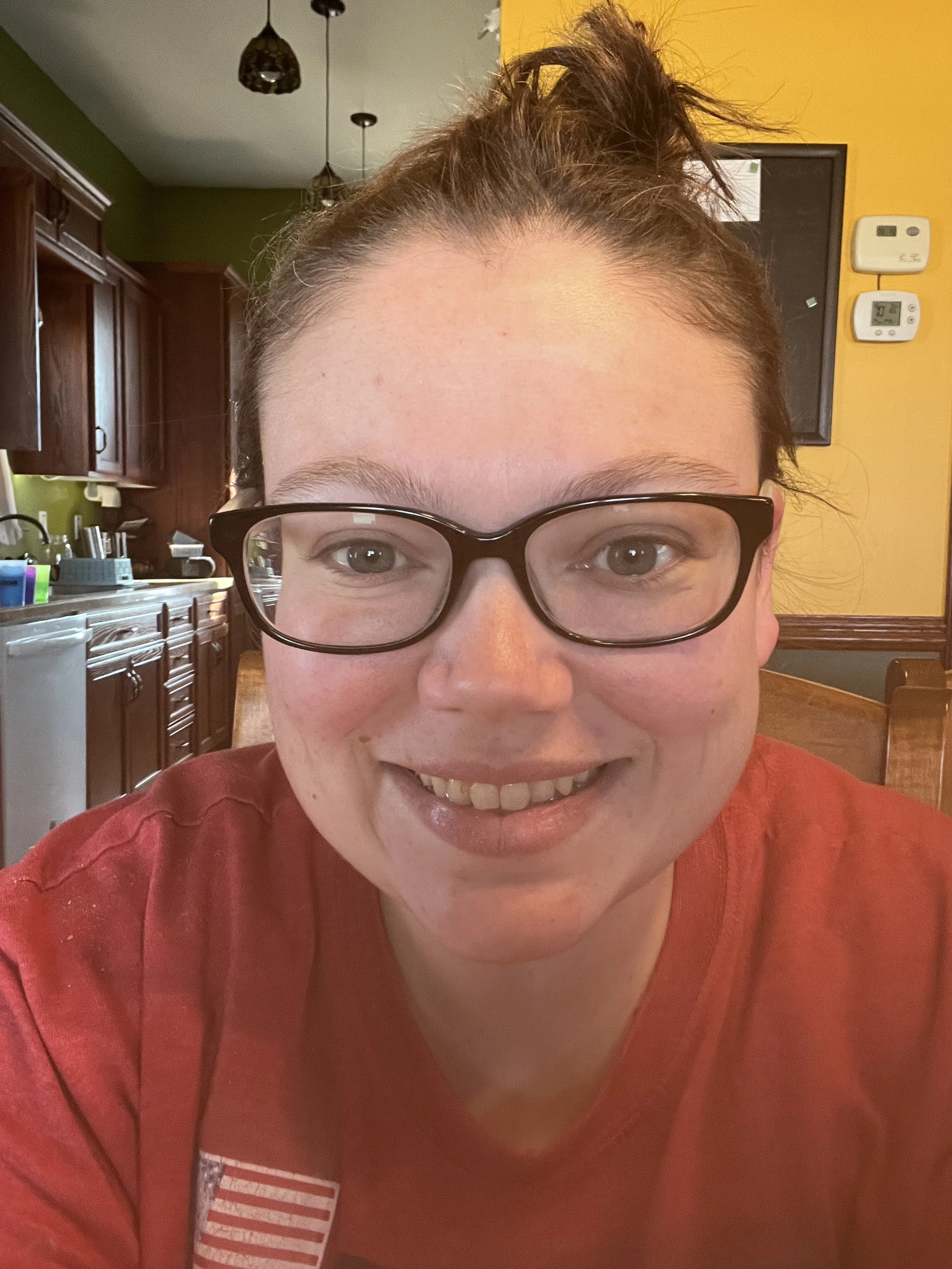 A person with glasses smiling, wearing a red shirt, in a kitchen with wooden cabinets and yellow walls.