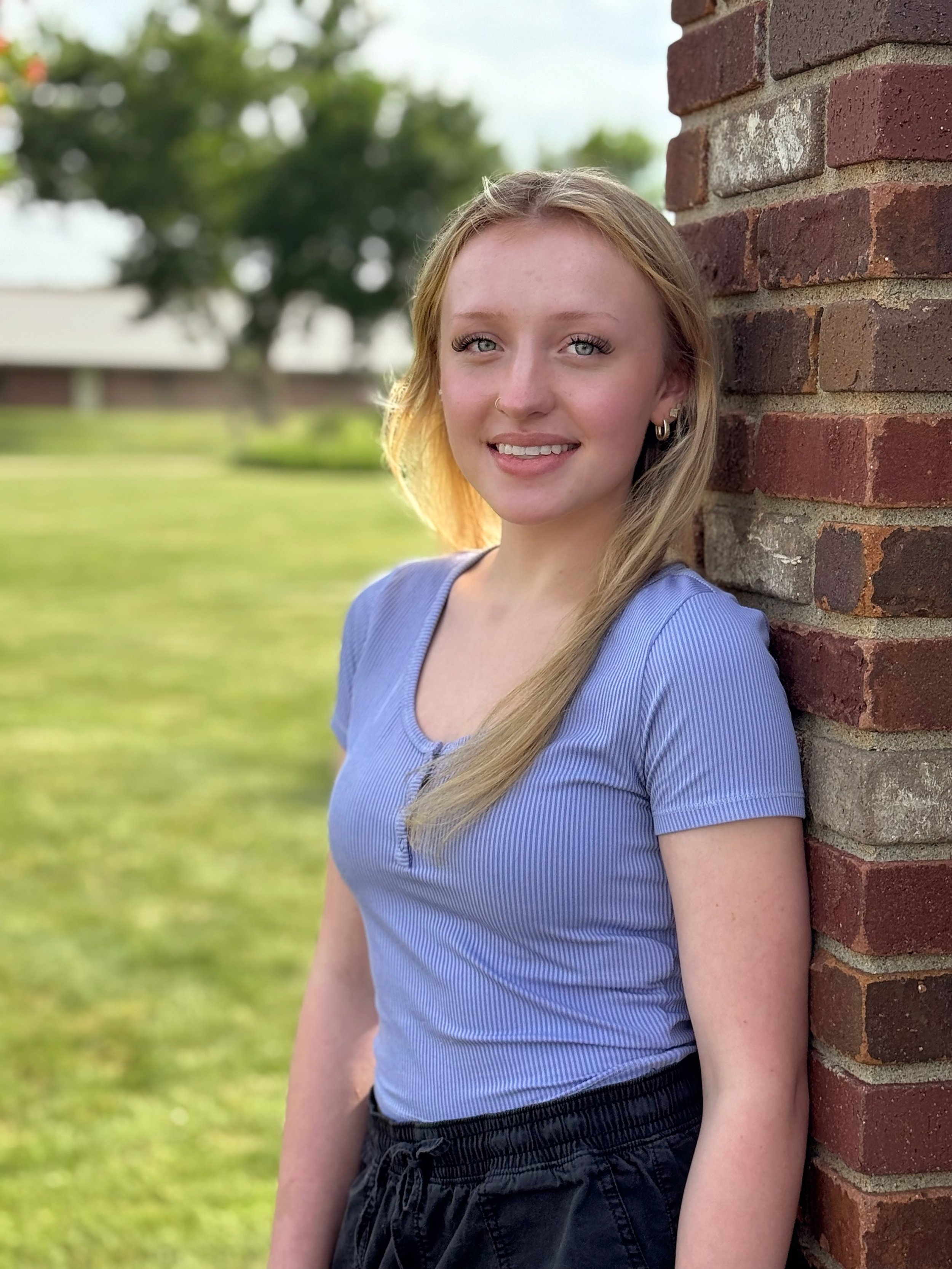 Young woman with blonde hair and blue eyes leaning against a brick wall outdoors on a sunny day, smiling at the camera.