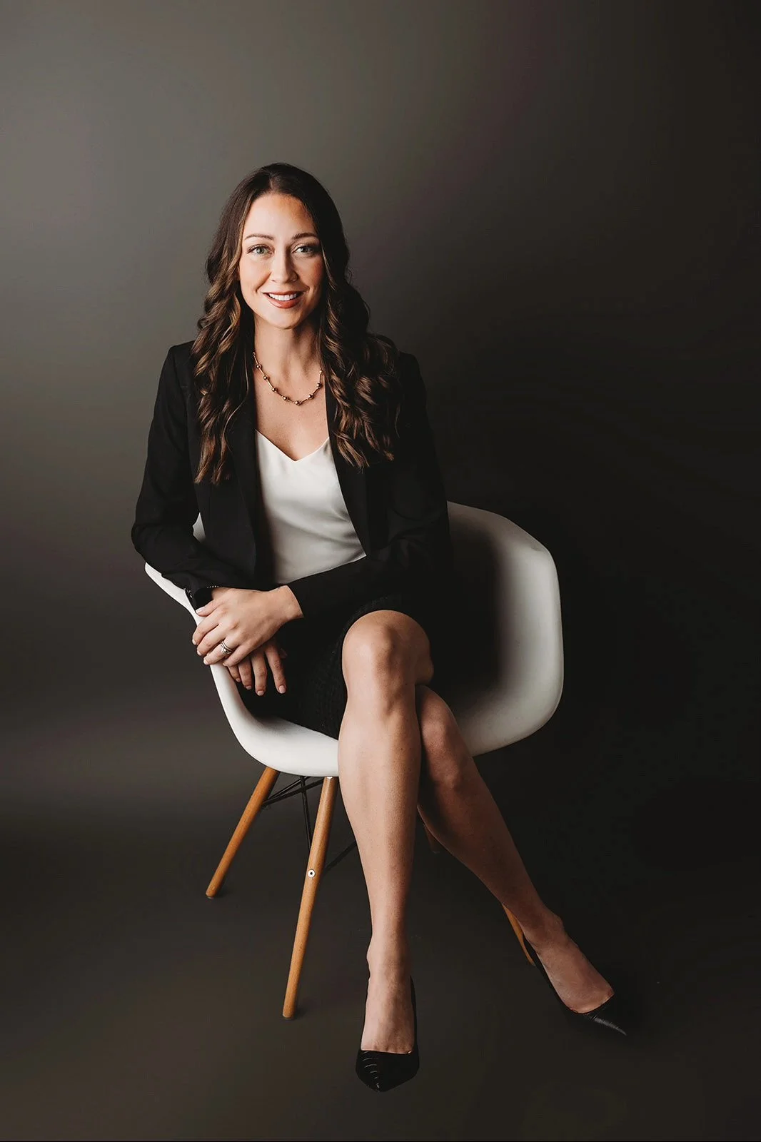 A woman sitting on a white chair with wooden legs, dressed in business attire, smiling at the camera against a dark background.