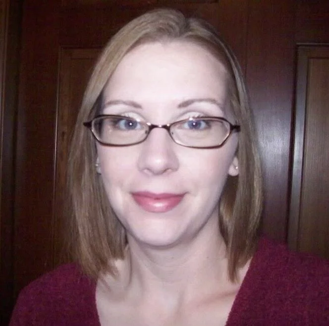 A woman with light skin and shoulder-length light brown hair wearing glasses and a maroon top in front of a wooden background.