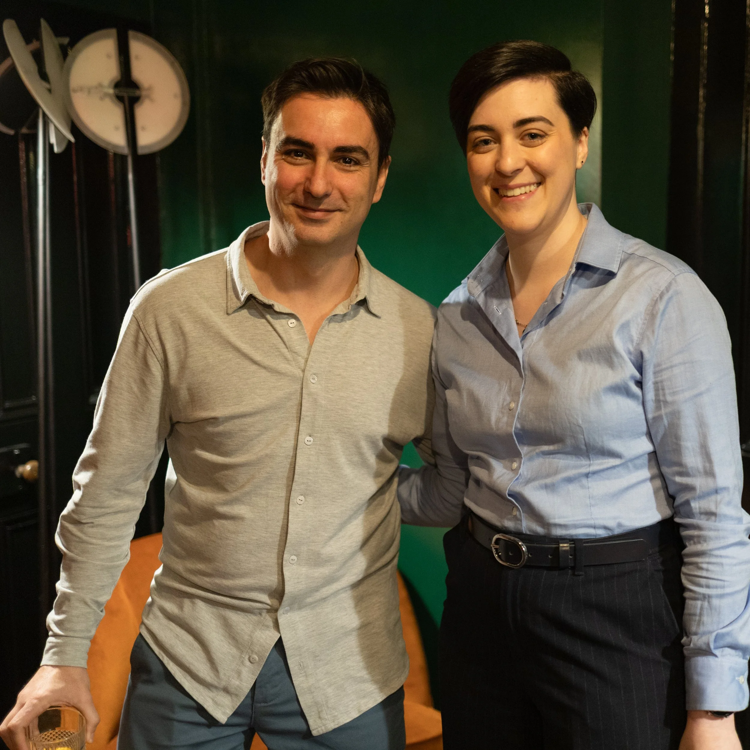 A man and woman smiling, standing close together in a room with a dark background, a green wall, and a clock in the background.