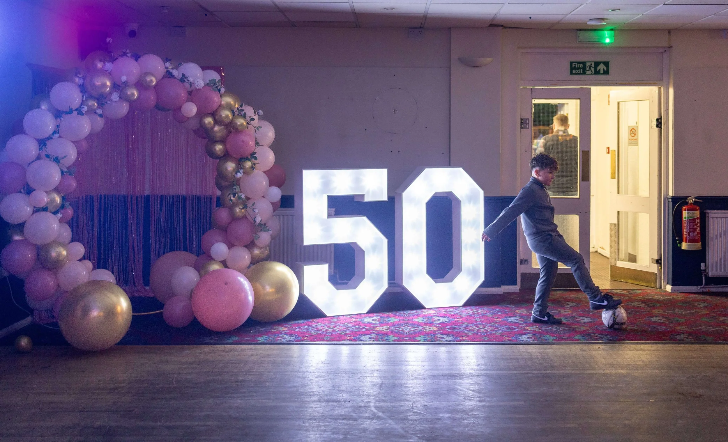 Young boy playing football in front of a brightly lit 50th birthday sign 
