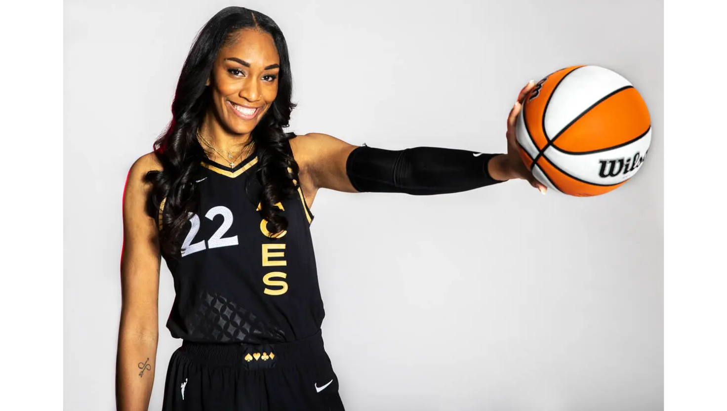 A young woman in a black basketball jersey holding a basketball, smiling and extending her arm toward the camera.