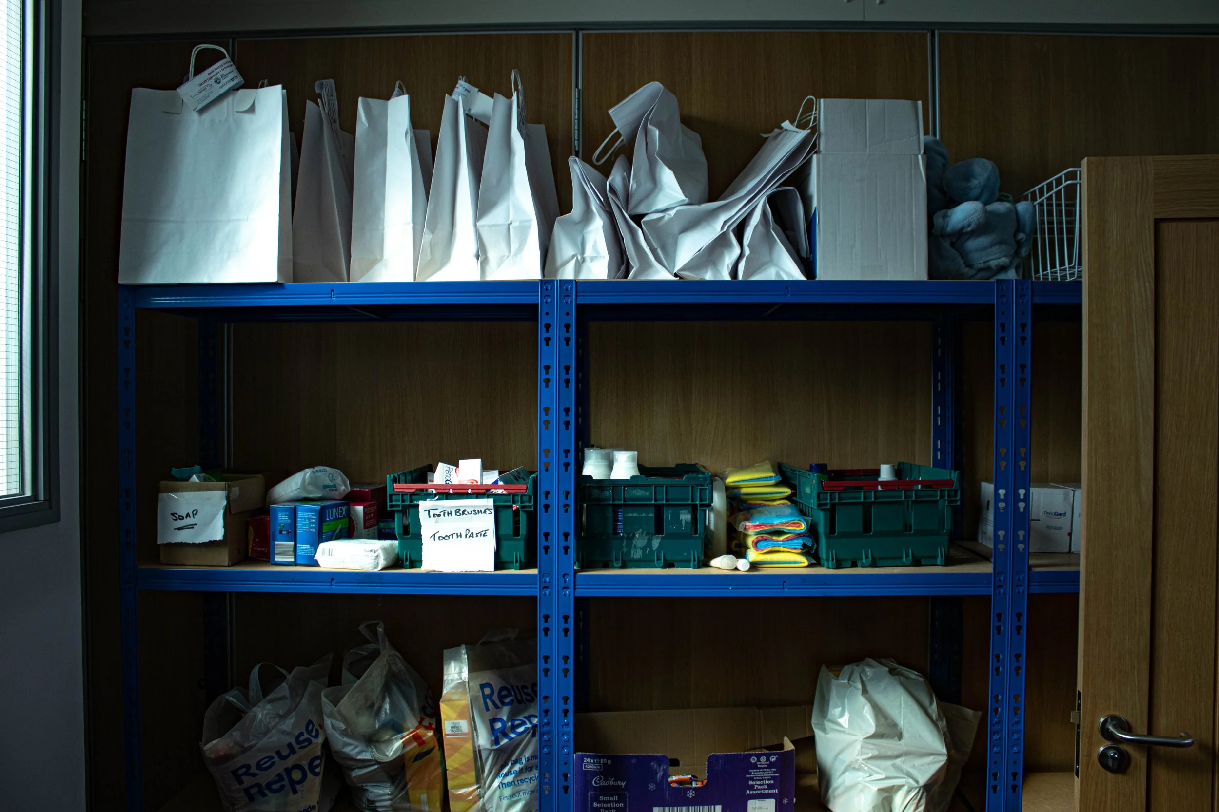 Shelving unit with paper bags and labeled boxes, containing toiletries like soap, toothbrushes, and toothpaste.