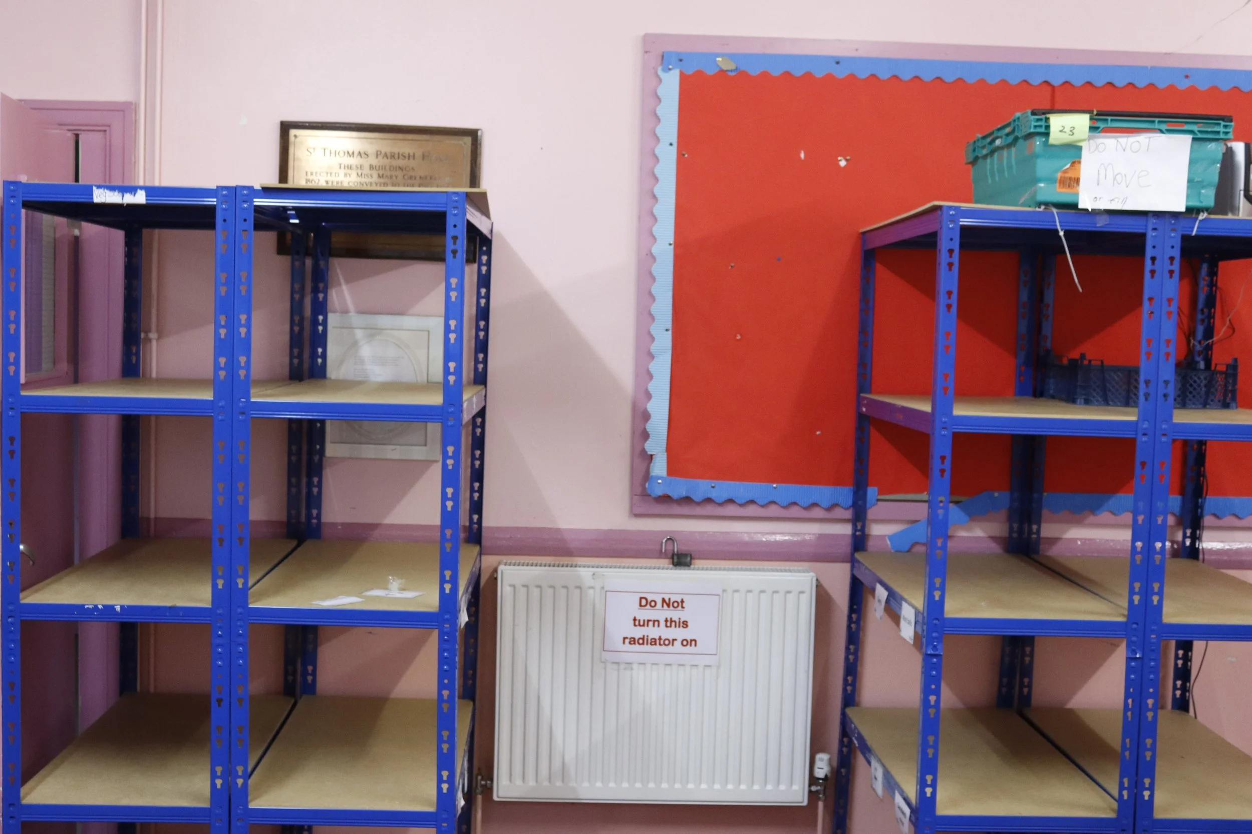 Empty blue metal shelving units against a light pink wall, adjacent to a radiator with a "Do Not turn this radiator on" sign. A bulletin board with a red background and decorative border is in the background, along with a green box labeled "Do Not Mo