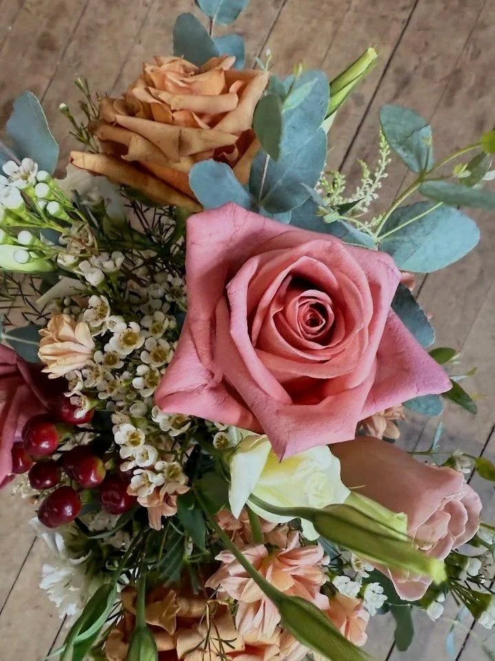 A bouquet of various flowers including a pink rose, a white rose, brownish roses, small white flowers, red berries, and green leaves on a wooden surface.