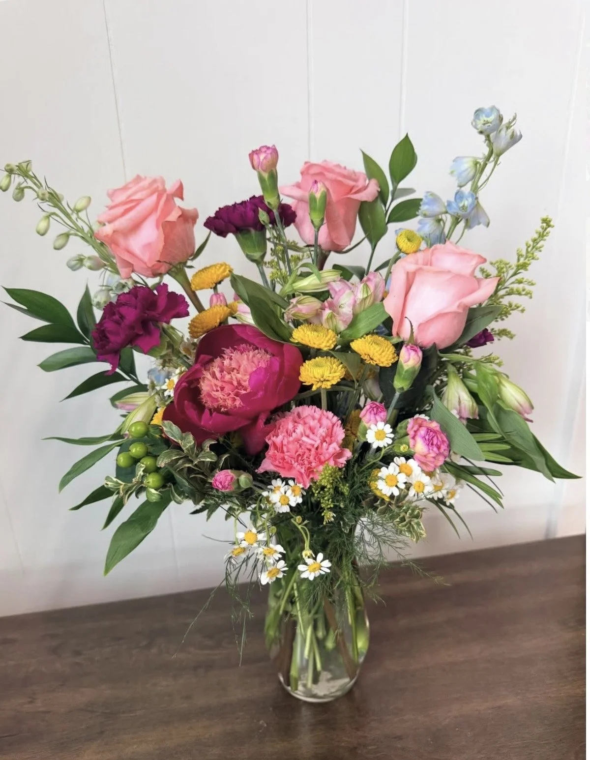 A bouquet of mixed flowers in a glass vase on a wooden surface features pink roses, purple and pink carnations, blue delphiniums, yellow daisies, and greenery.