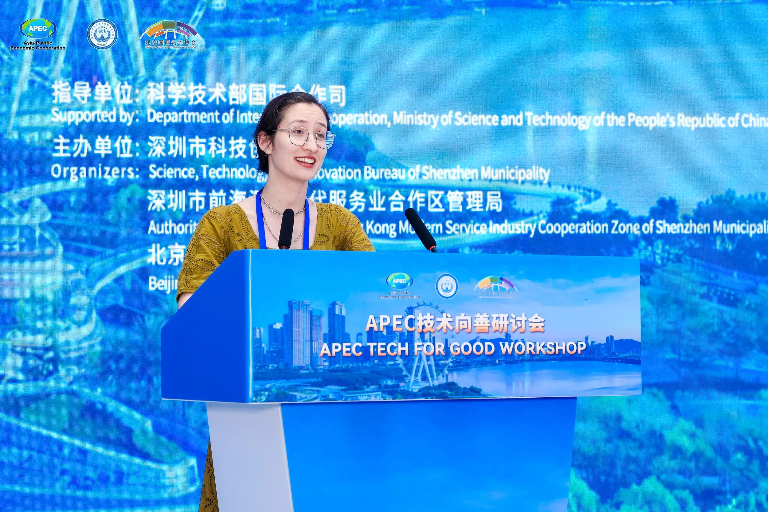 A woman speaking at the APEC Tech for Good workshop in Shenzhen with a blue backdrop displaying logos and text in Chinese and English.
