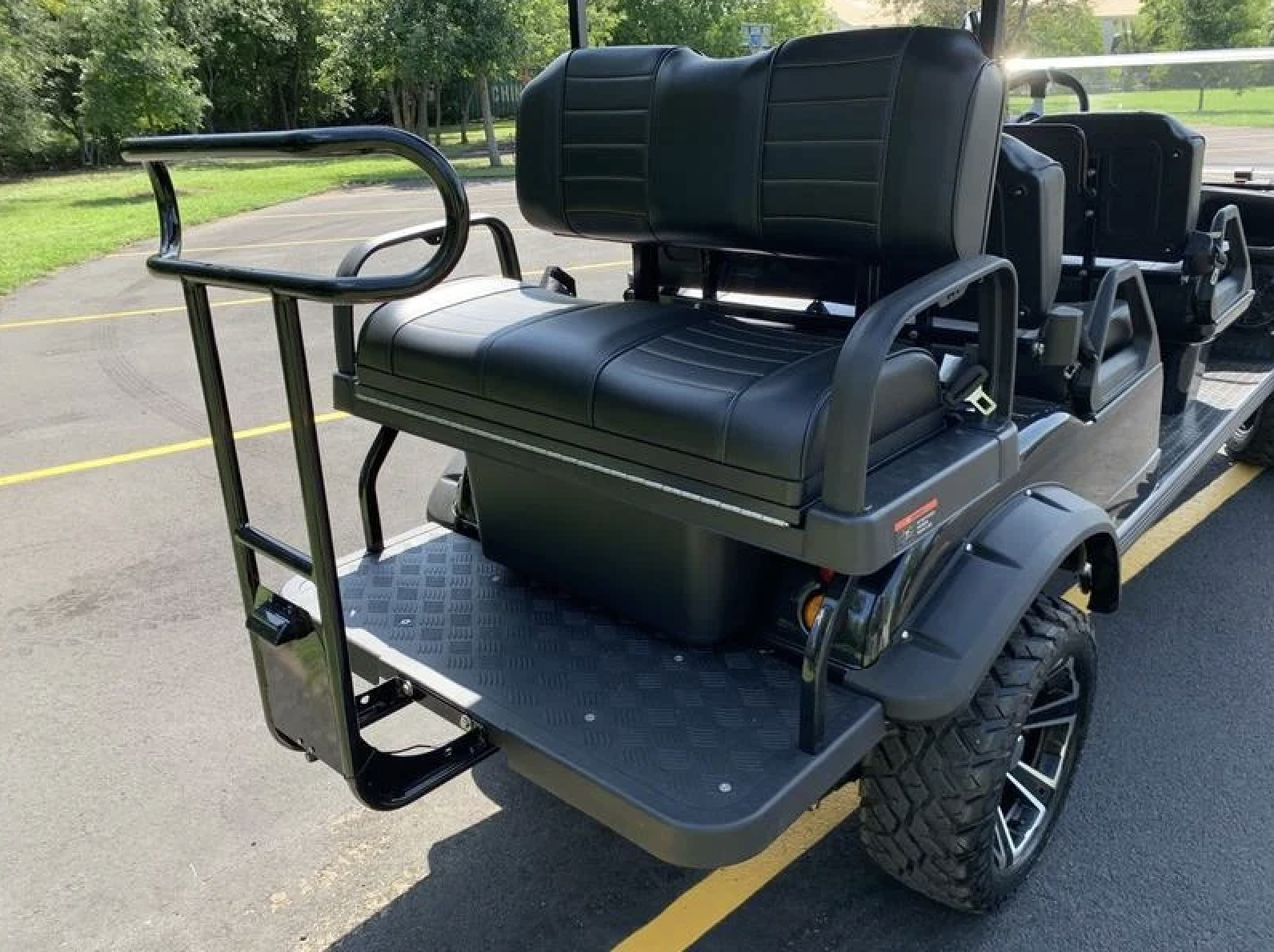 Black utility cart or vehicle with seating and a luggage rack, parked on asphalt with parking lines, surrounded by green trees and grass.