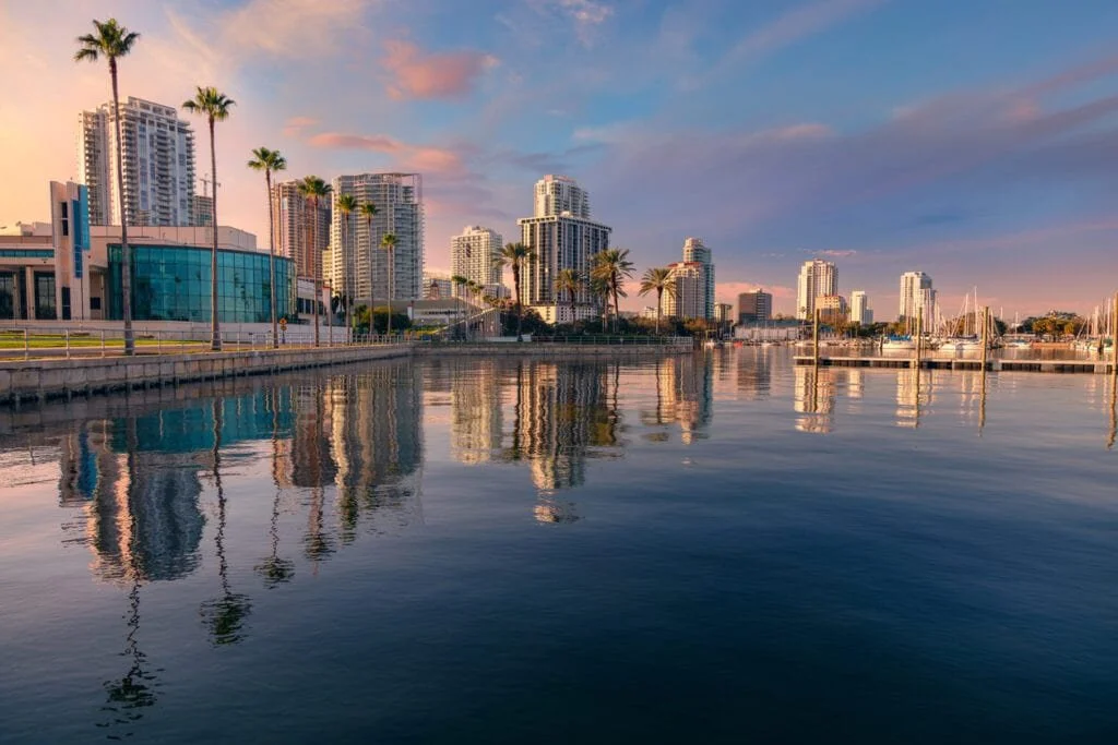 Sunset over the downtown St. Petersburg, Florida waterfront with palm trees, reflections, and city skyline glowing in golden hour light.