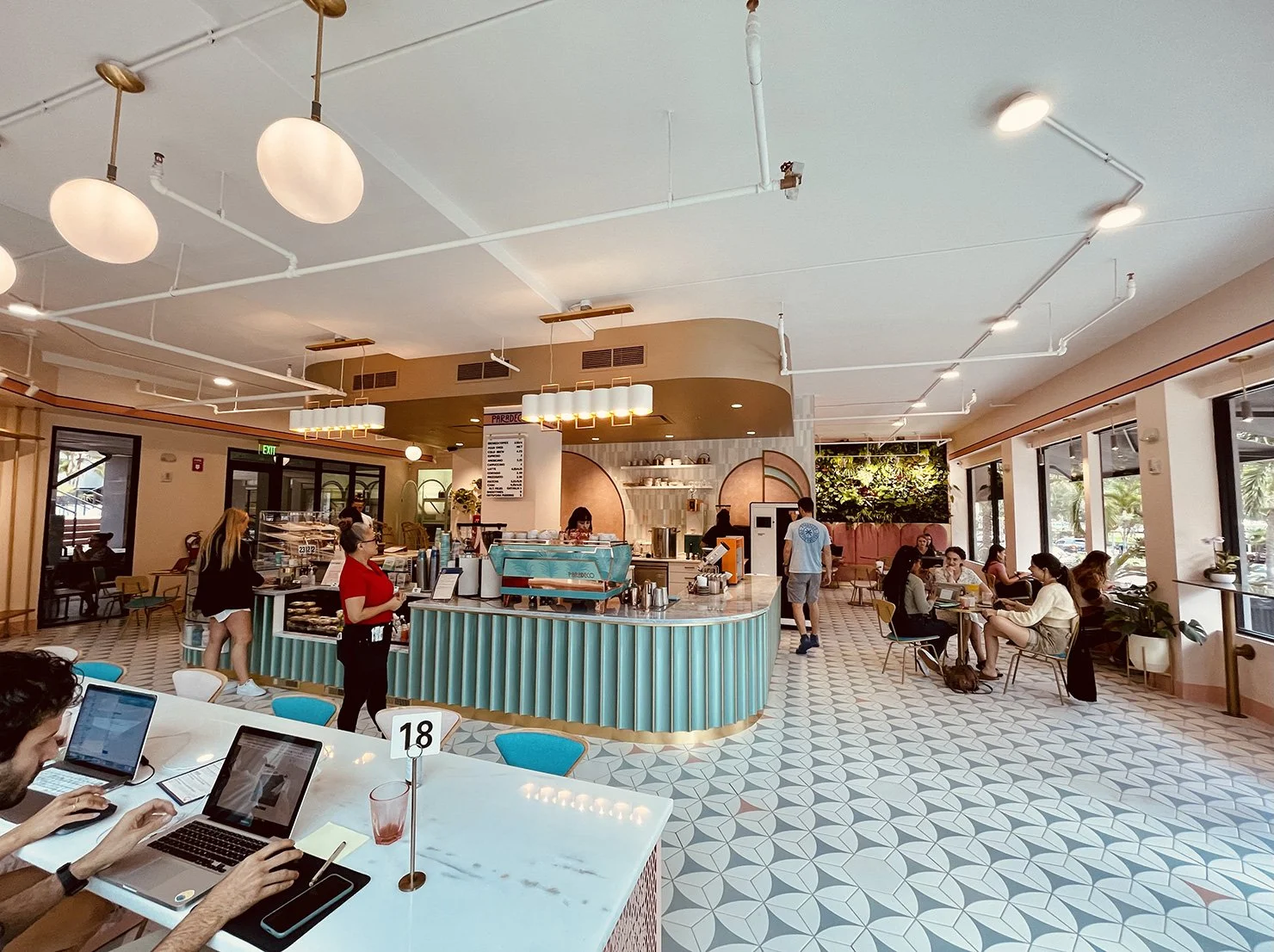 Interior of Paradeco Coffee in St. Pete featuring pastel decor, patterned tile floors, modern lighting, and customers working and enjoying their drinks.