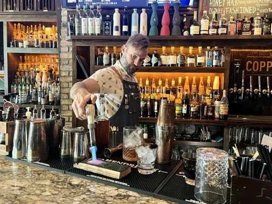 Bartender crafting a cocktail at Copper Shaker in downtown St. Pete, surrounded by warm lighting and shelves of spirits.