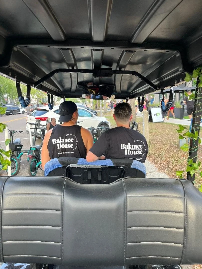 Two people wearing Balance House shirts riding in a street-legal golf cart in downtown St. Petersburg, Florida.
