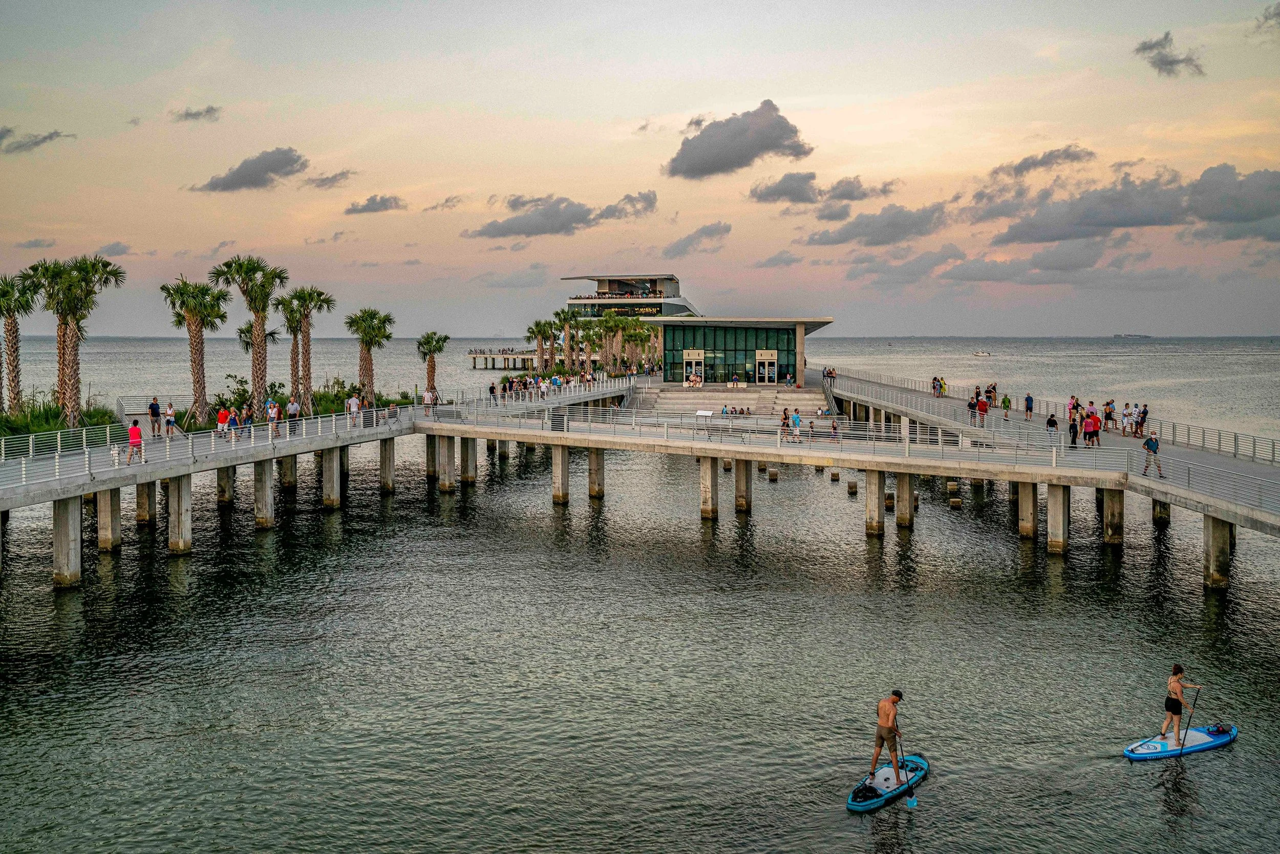 People walking along the St. Pete Pier during golden hour with paddleboarders on the water and soft sunset colors in the sky.