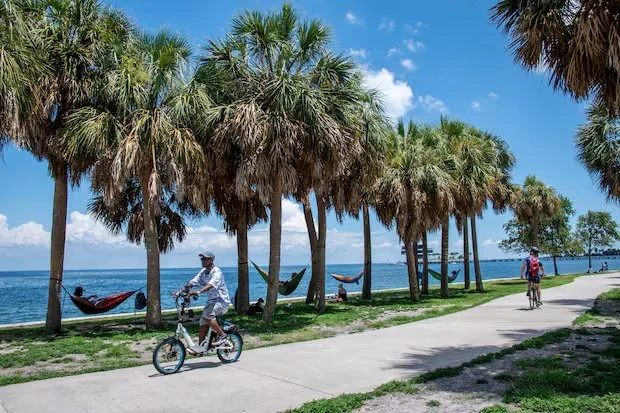 People relaxing in a hammock under shady trees at North Shore Park in St. Pete, overlooking the waterfront on a sunny morning.