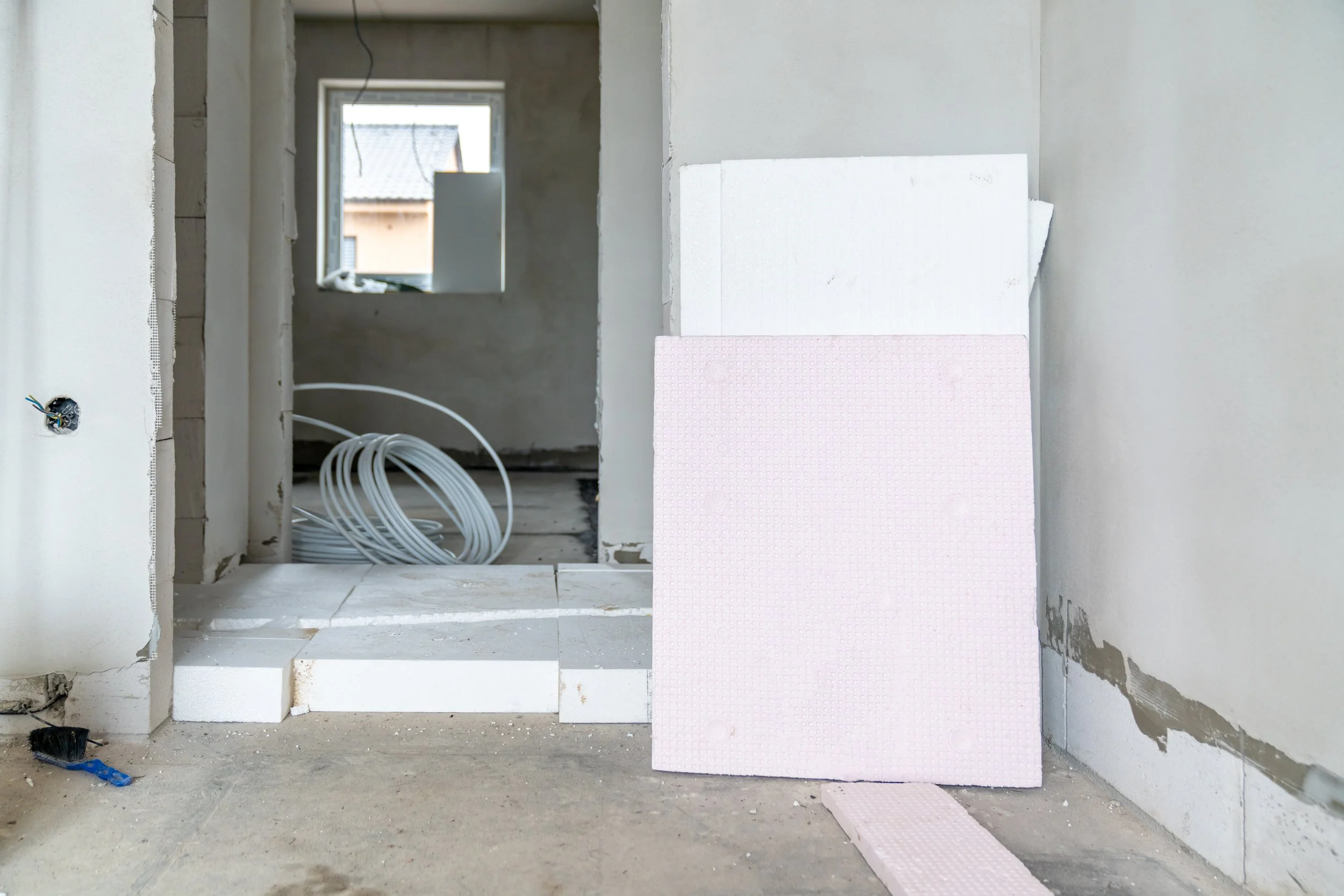 Interior of a room under construction with a doorway, showing electrical wiring on the floor, a window in the background, and insulation boards leaning against the wall.