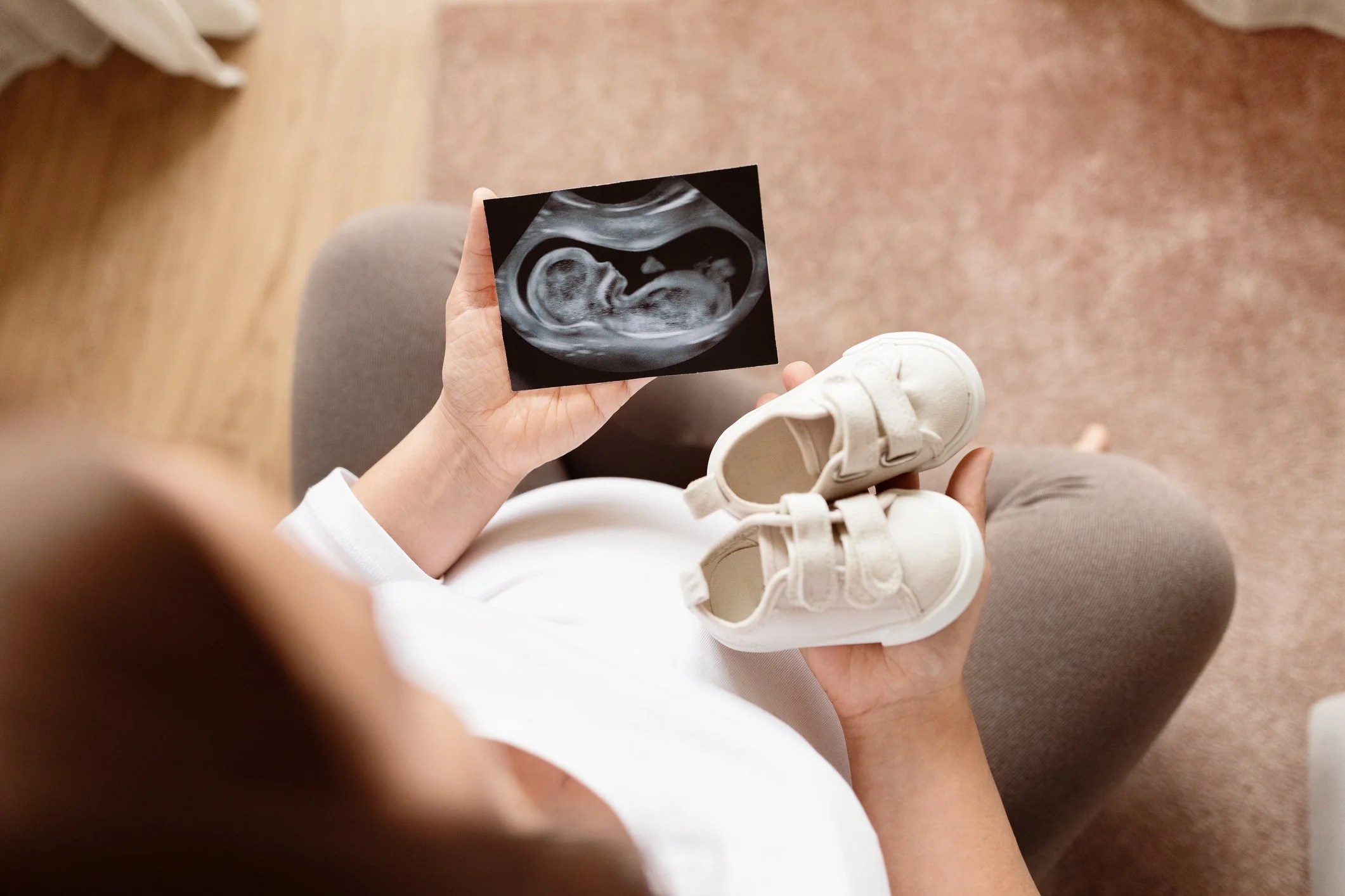 A pregnant woman sitting on a chair holding a sonogram of her developing baby, along with a pair of white baby shoes wondering what the birth will be like.