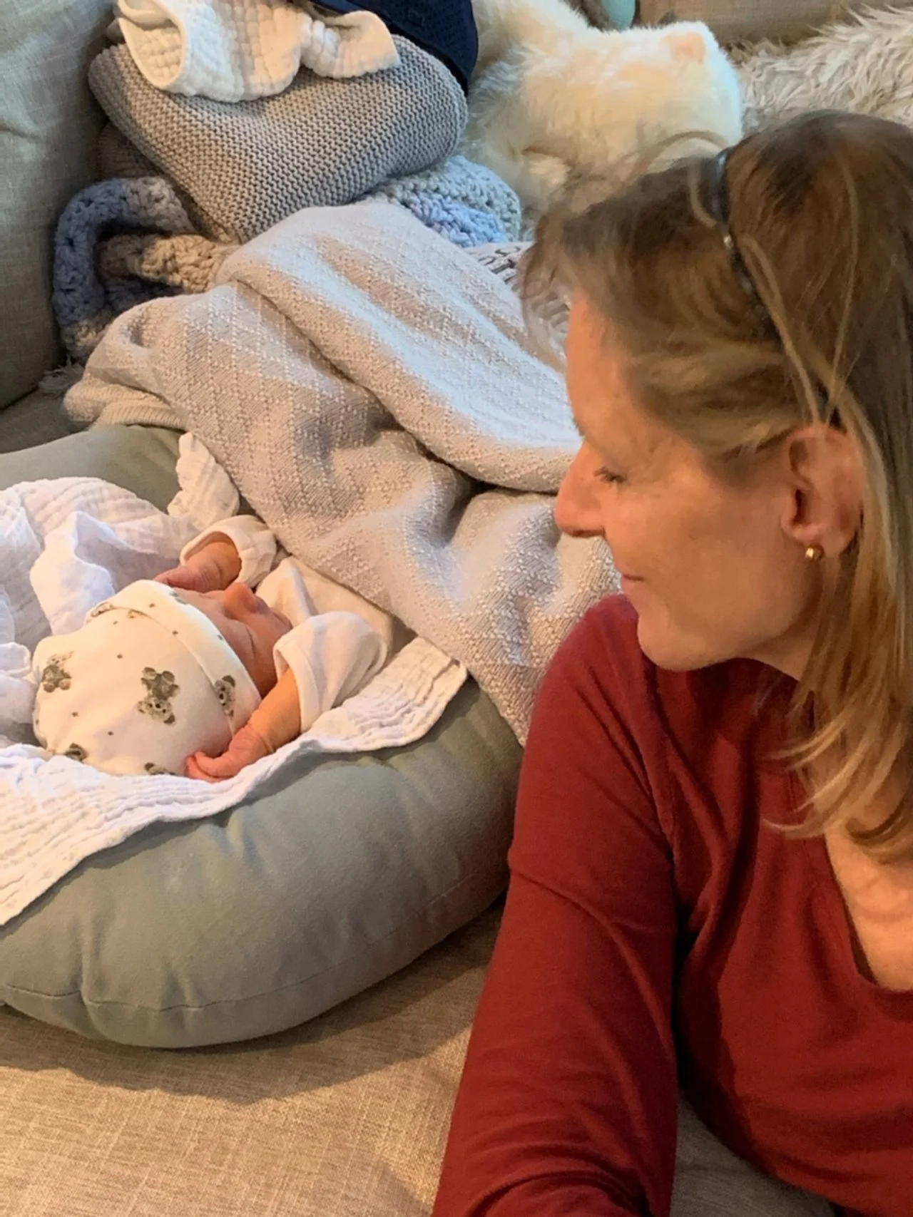 A caring and skilled woman looking at a sleeping newborn baby on a cushioned baby lounger surrounded by blankets and stuffed animals, with a small dog sleeping nearby.