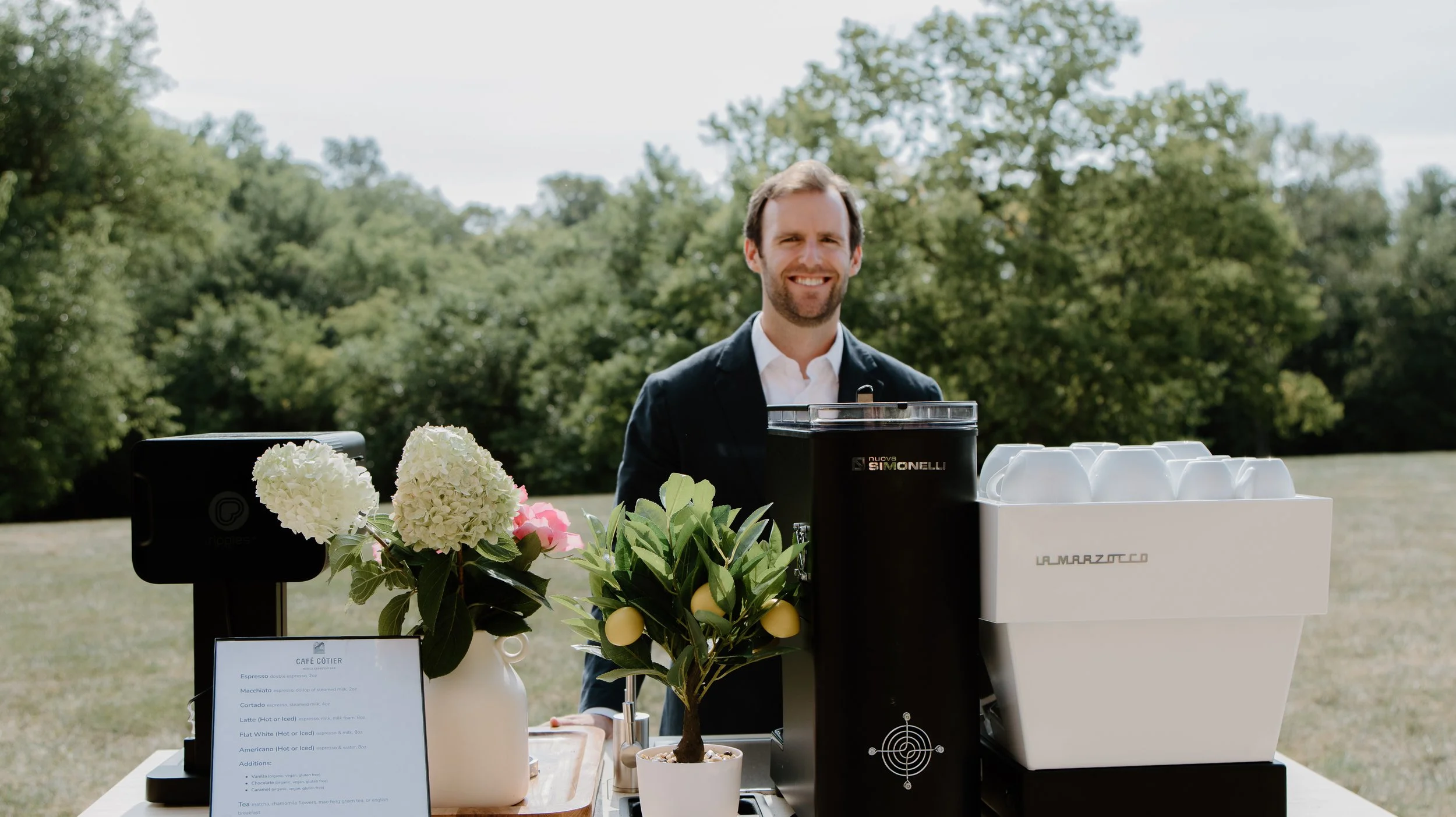 Smiling man standing behind a coffee station outdoors with a black coffee machine, white cups, a flower arrangement, and a small lemon tree
