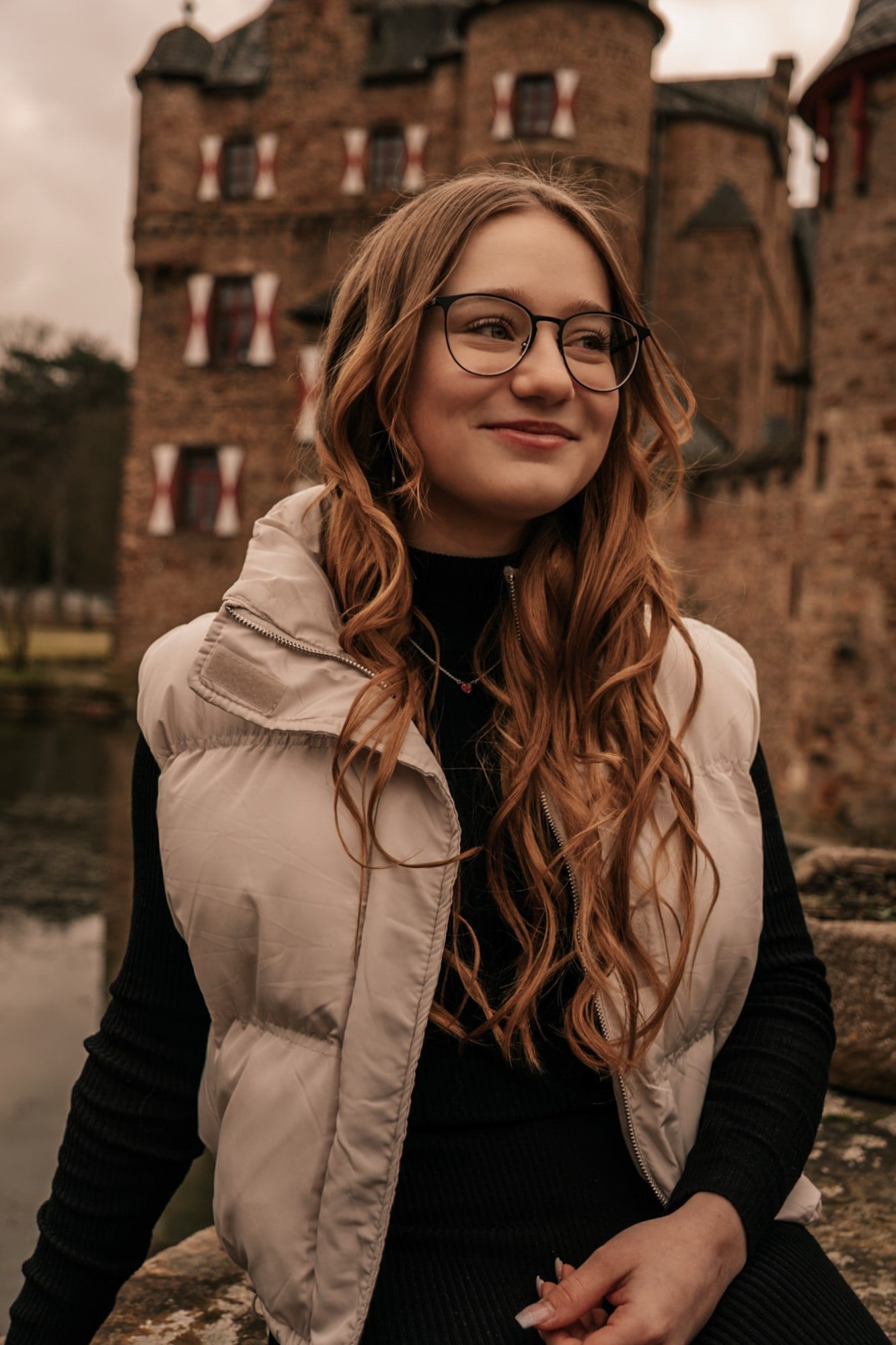 Junge Frau mit Brille und langer, lockiger Haaren, trägt eine helle Weste vor einem alten, steinernen Gebäude mit mittelalterlichem Erscheinungsbild.