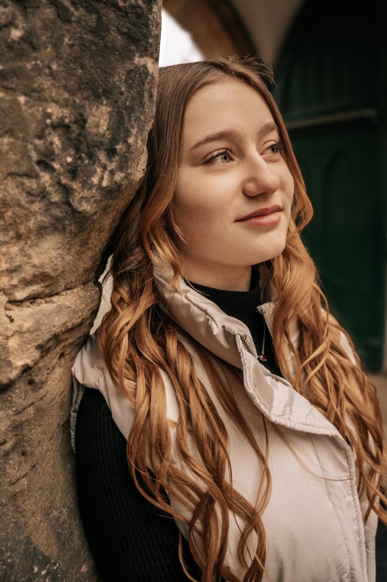 Nahaufnahme einer Frau mit langen, lockigen Haaren, die an einer Steinmauer lehnt und nachdenklich zur Seite schaut. Sie trägt eine helle, wattierte Weste und einen schwarzen Pullover.