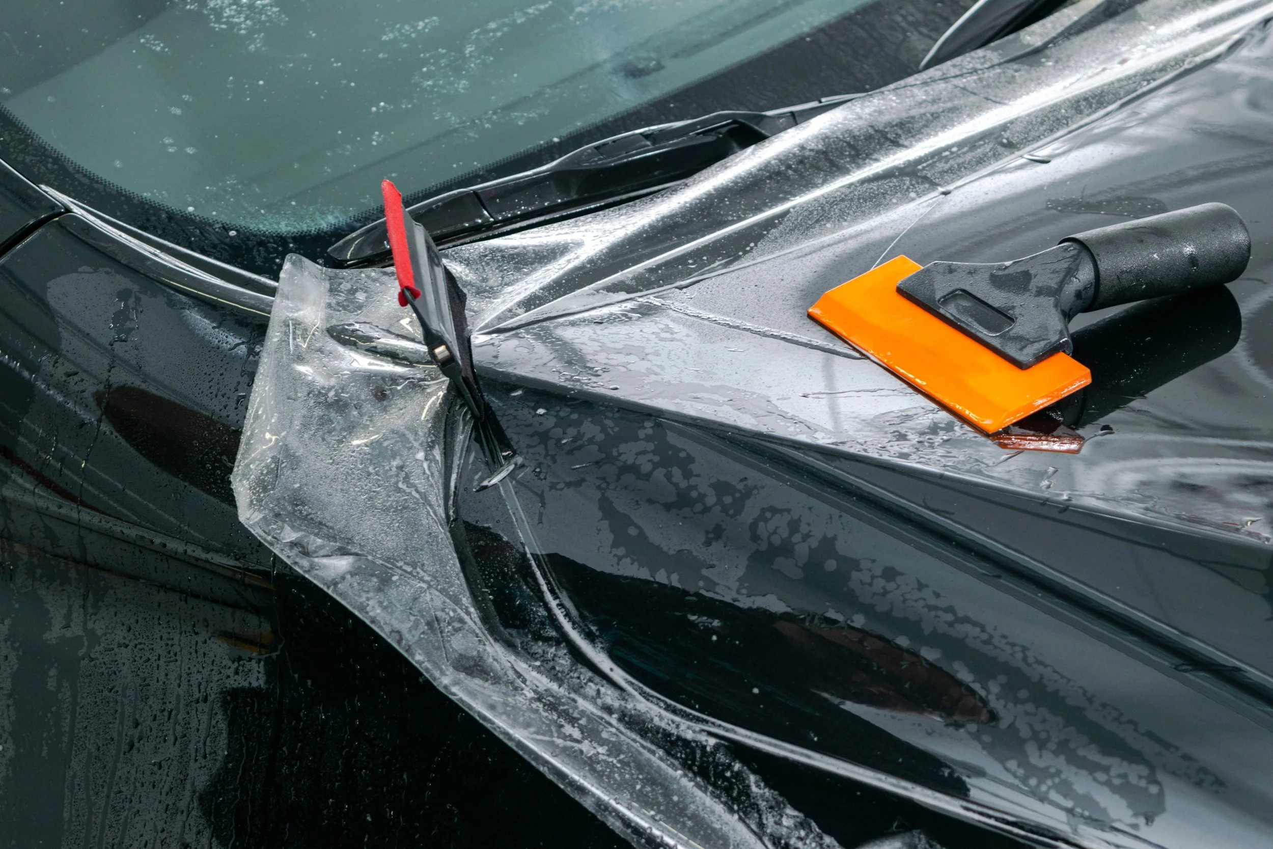 Broken windshield of a black car with a plastic squeegee and a scraper on the hood, indicating a car accident or damage.