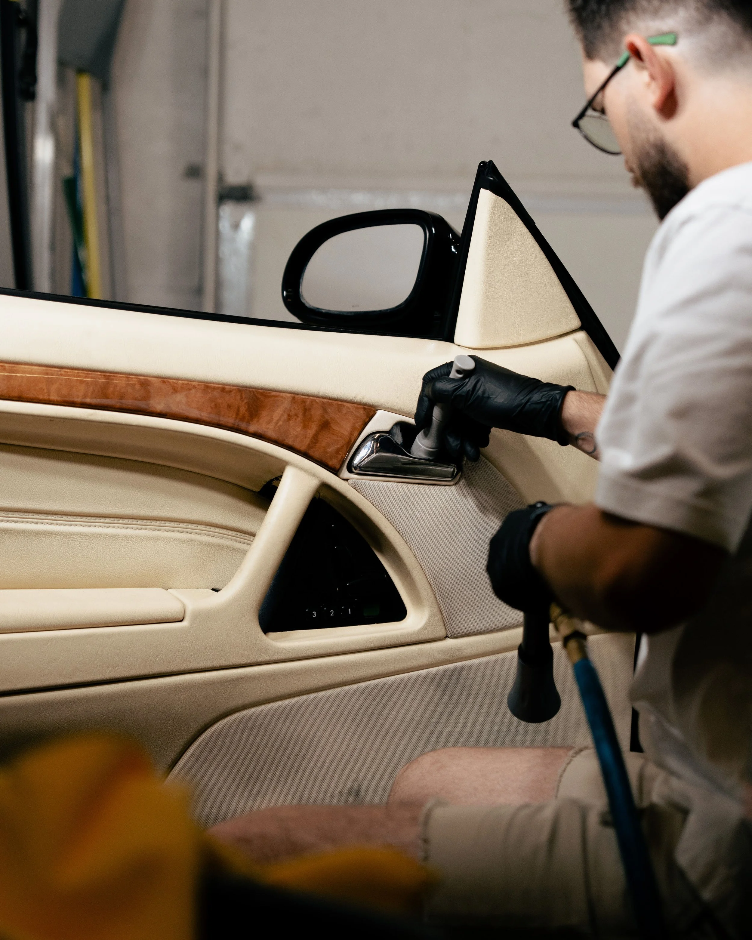 A person wearing glasses and black gloves is cleaning the dashboard of a classic beige car with a wood accent in a garage.