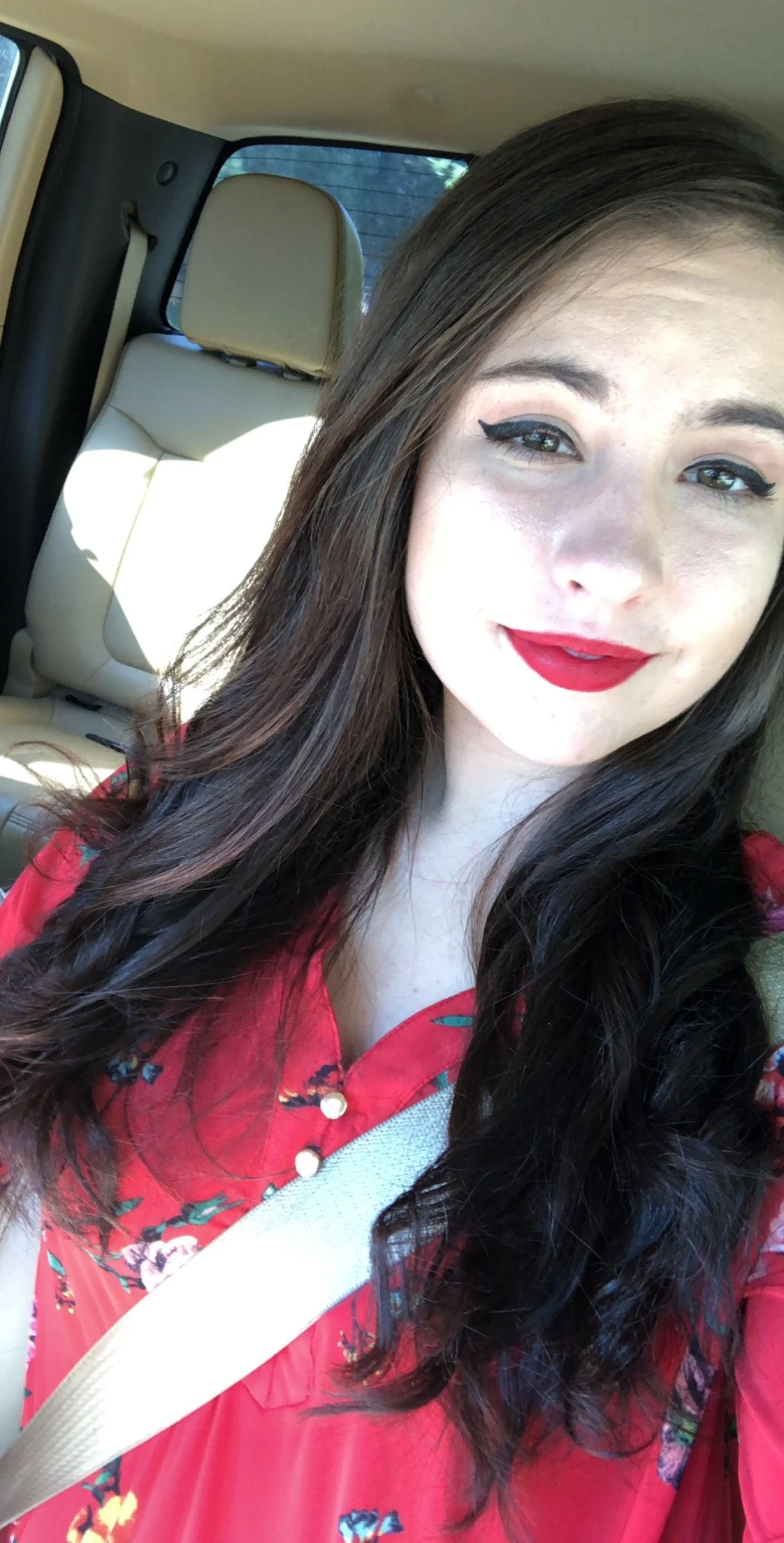 A young woman with long dark hair, wearing red lipstick and black eyeliner, taking a selfie inside a car with beige interior.