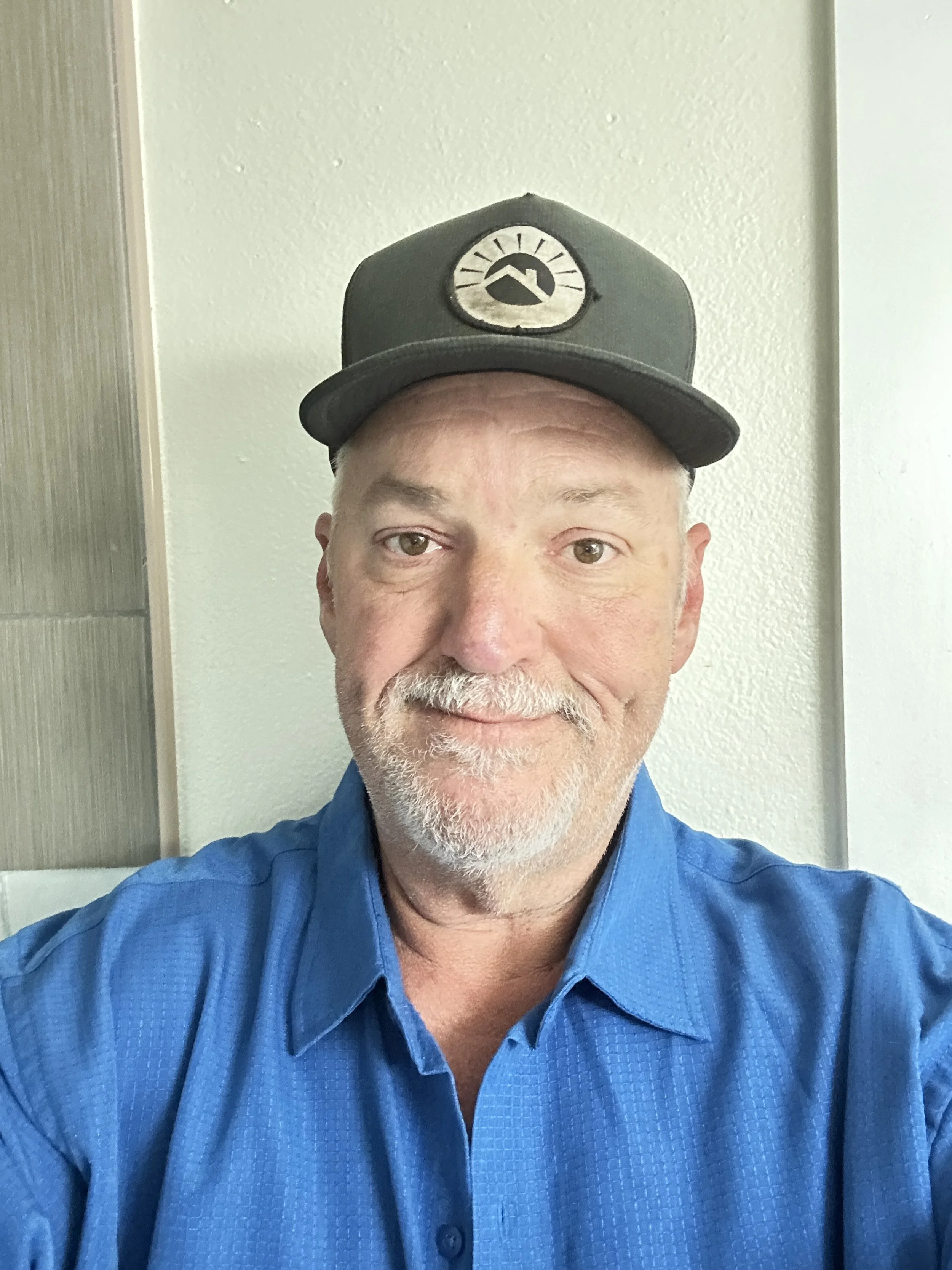 A middle-aged man with a beard and mustache wearing a blue collared shirt and a black baseball cap with a mountain logo, smiling for a selfie against a light-colored wall.
