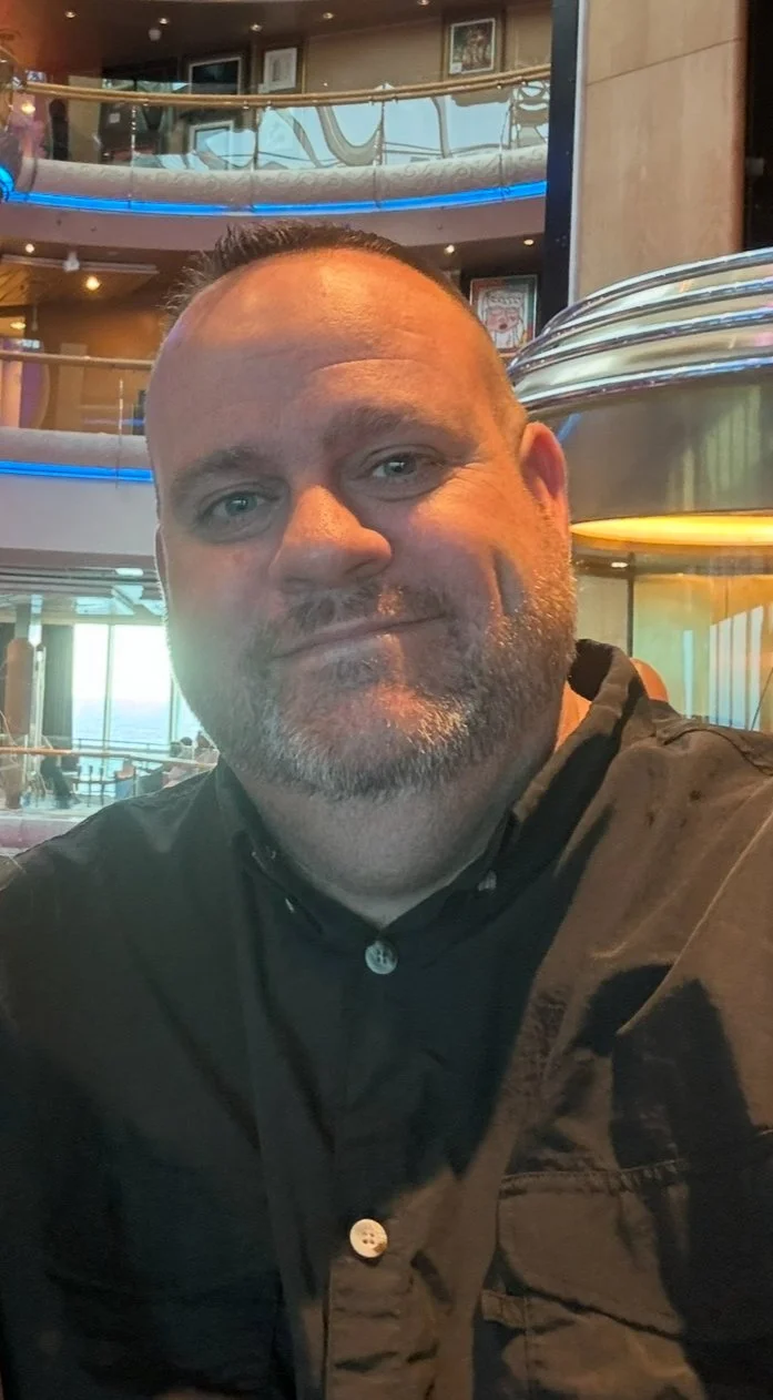 A man with a beard and short hair smiling in an indoor setting, possibly a cruise ship or a resort, with multiple levels and large windows in the background.