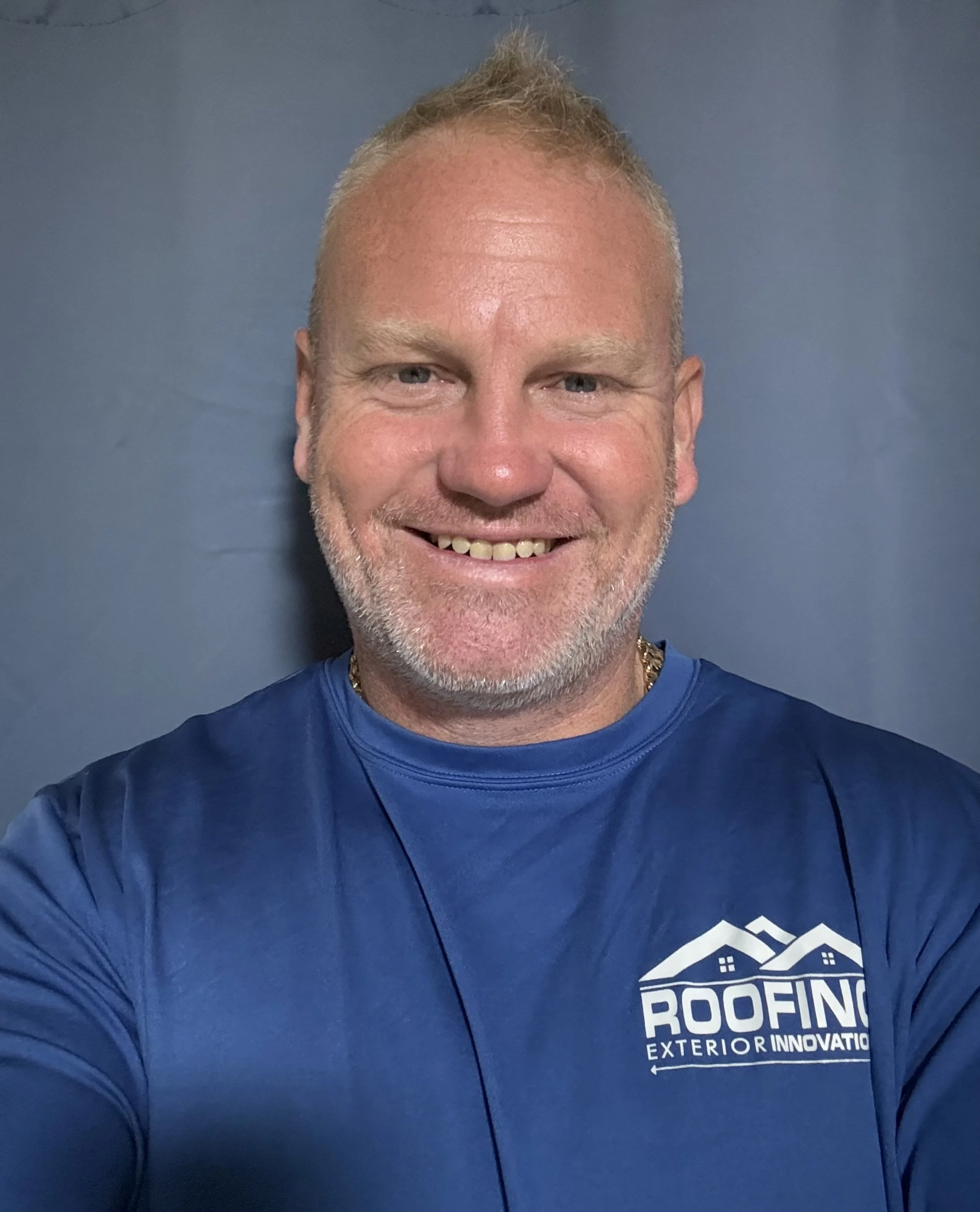 A smiling man with short blond hair and a beard wearing a blue T-shirt with a roofing company logo, standing against a plain blue background.