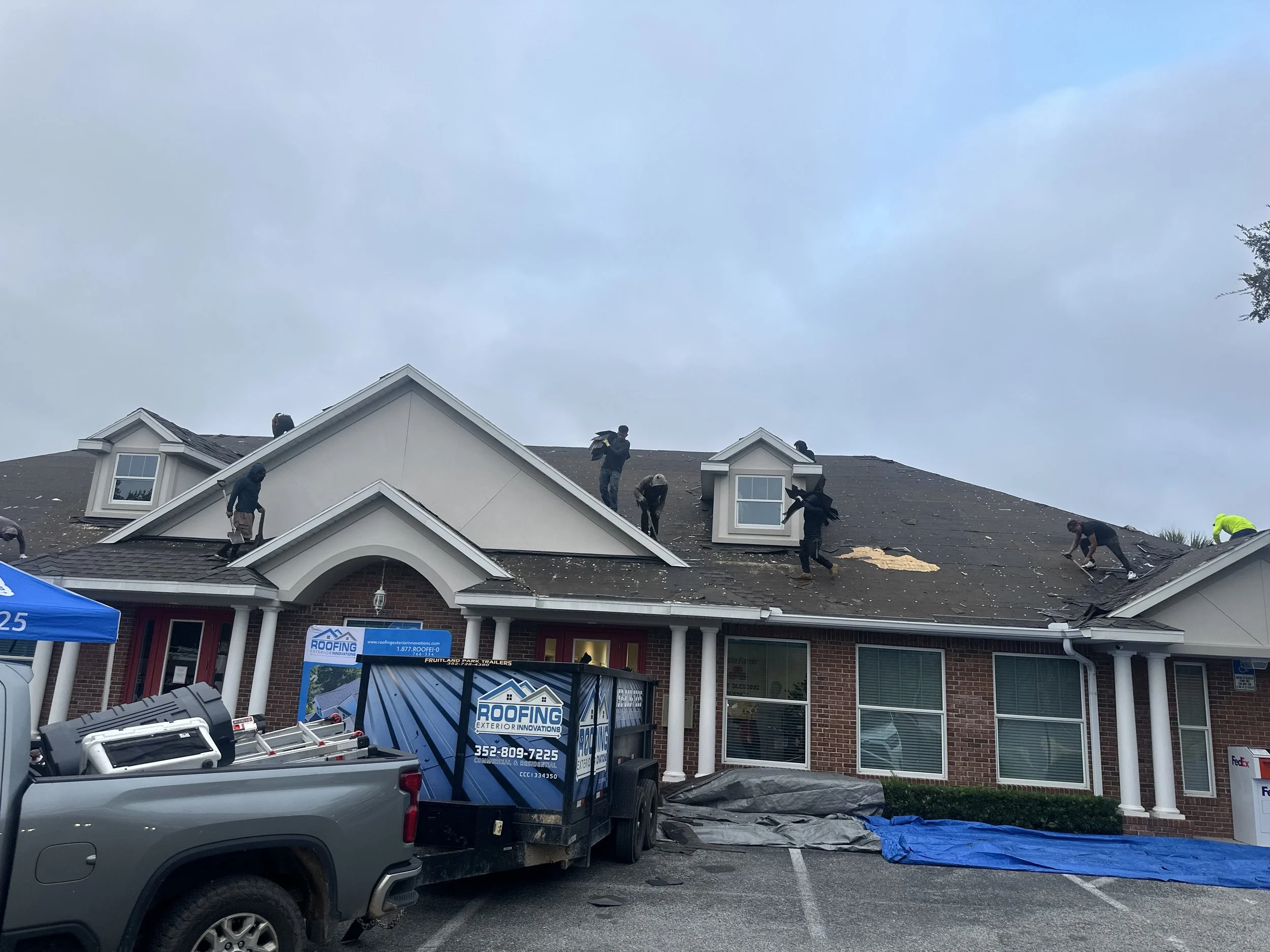 Workers repairing or replacing shingles on the roof of a brick commercial building with a parking lot in front, cloudy sky overhead, and roofing equipment nearby.