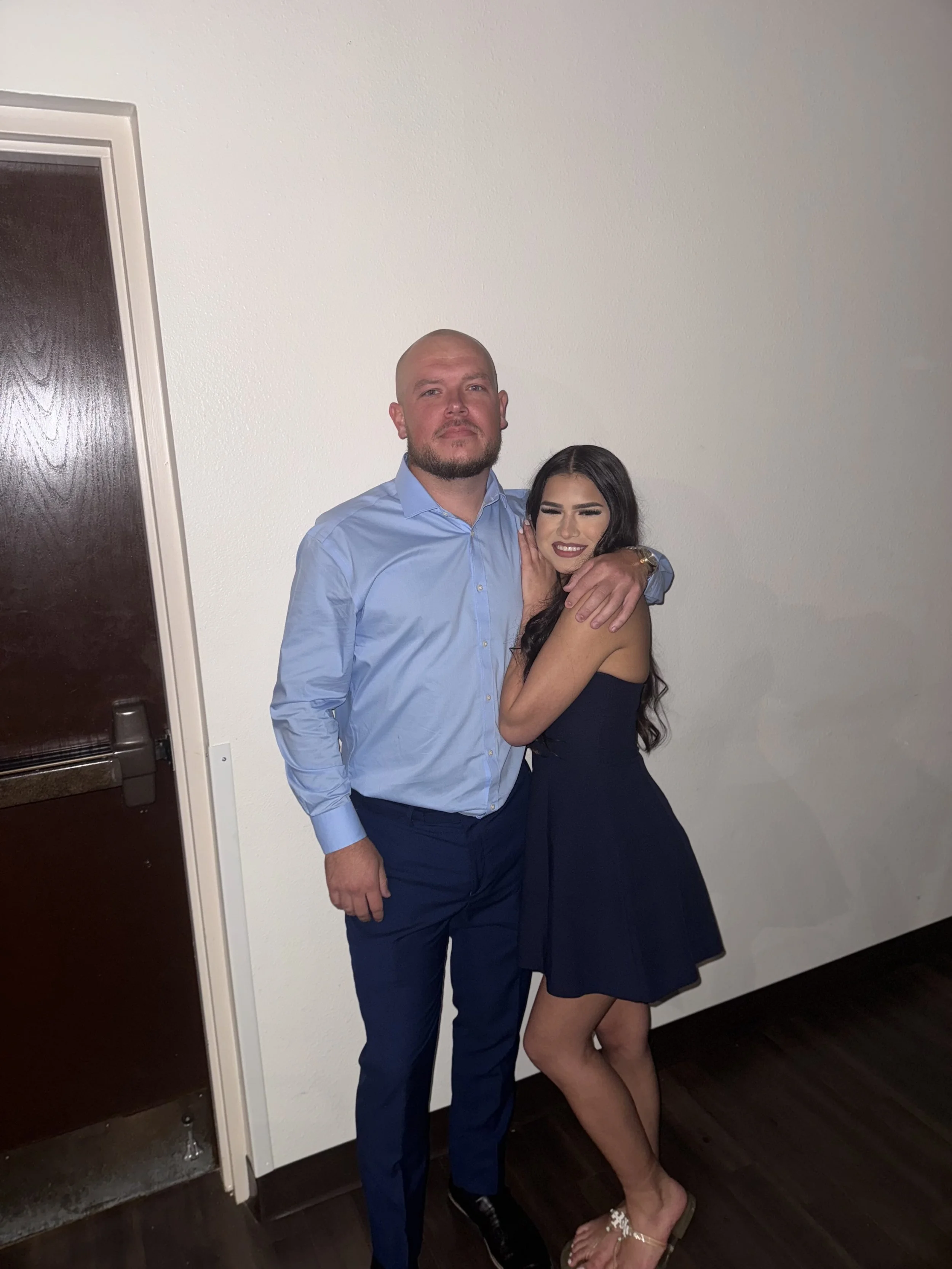 A man in a light blue shirt and dark blue pants standing with a woman in a navy dress, hugging her, both smiling, in front of a plain white wall.