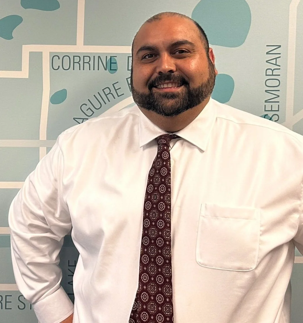 A man with a beard and shaved head smiling, wearing a white dress shirt and a patterned tie, standing in front of a background with city map designs and street names.