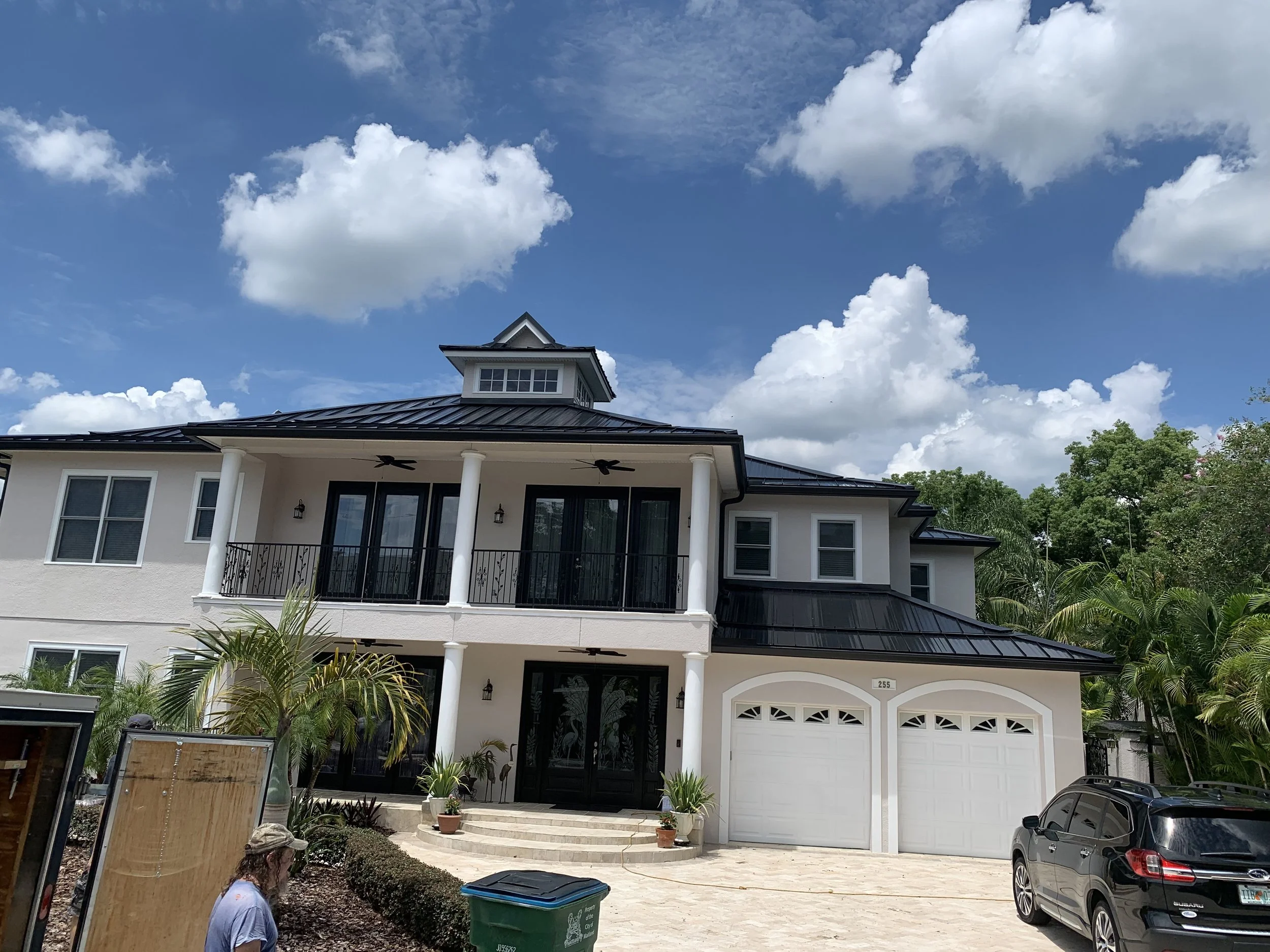 Large modern two-story house with white exterior, black roof, and a double garage, surrounded by tropical landscaping and parked black car, under a partly cloudy sky.