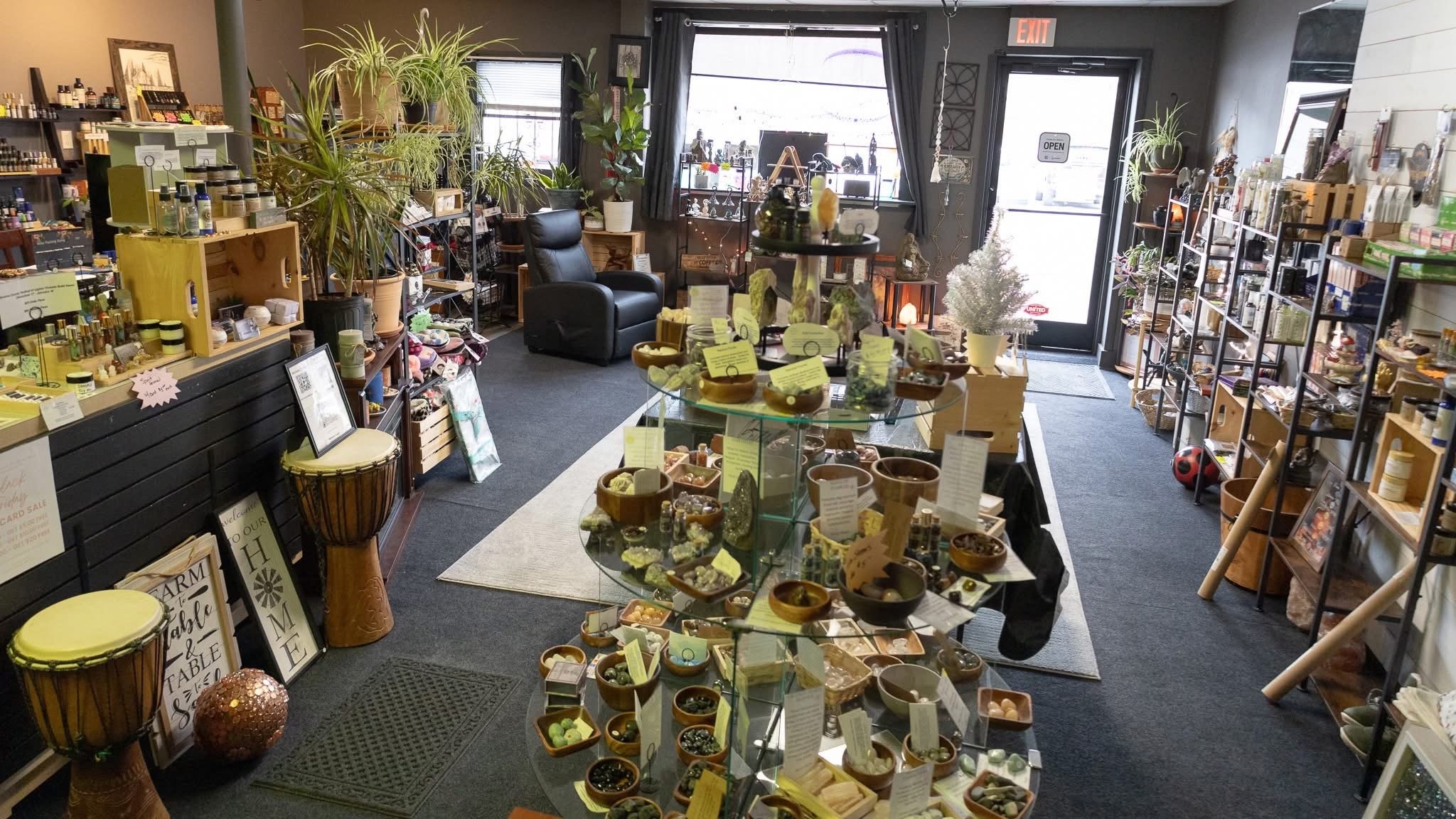 Interior of a gift shop with display shelves and tables filled with crystals, candles, plants, and decorative items, including a small Christmas tree near the entrance.
