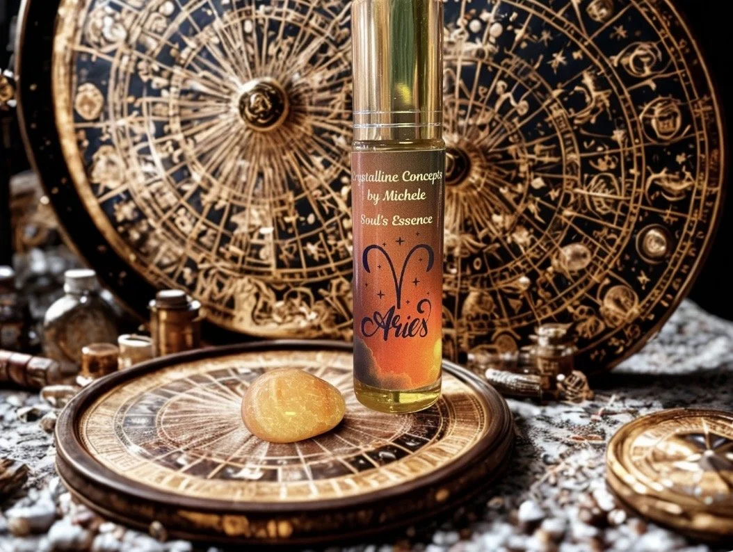 A bottle labeled 'Crystalline Concepts by Michele' and 'Soul's Essence' with 'Aries' zodiac symbol on it, placed on a round decorative surface resembling an astrological chart, with an orange sphere or gemstone in front of it. Behind, there are large, gold astrological wheels with zodiac symbols, surrounded by small vials and jewelry, all set on a textured surface.