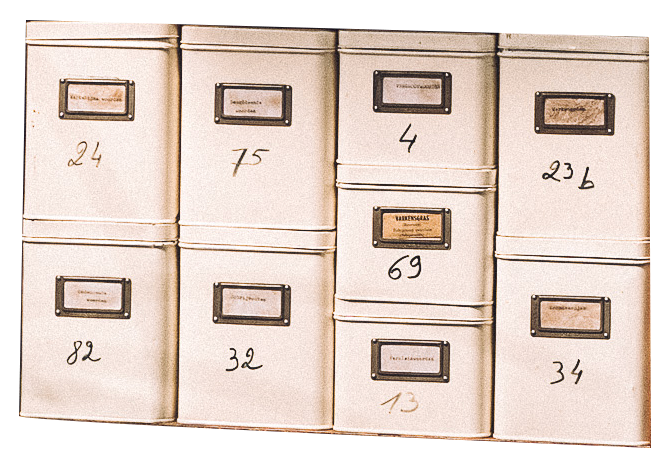 Wit metalen postvakken met handgeschreven nummers en enkele labels, gestapeld op een muur.