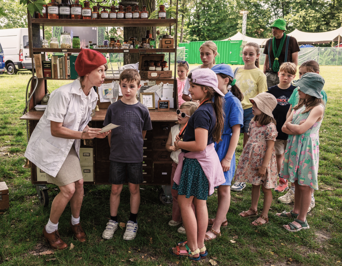 Een vrouw in een witte blouse en rode hoed spreekt met een groep kinderen die in een rij staan, in een park met een houten kraam gevuld met oude voorwerpen en boeken.