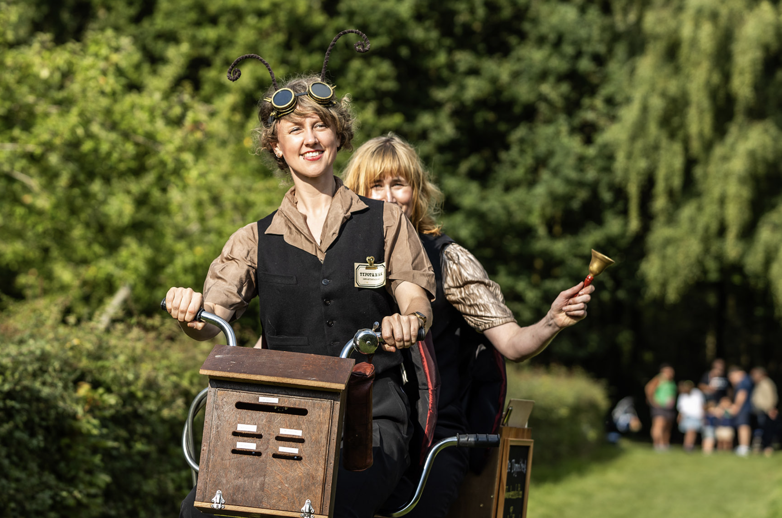 Twee vrouwen in middeleeuwse kleding rijden op een fiets met een houten doosje vooraan, één draagt opvallende antenne-achtige accessoires en een badge, de ander houdt een bel, alles in een groene buitenomgeving.