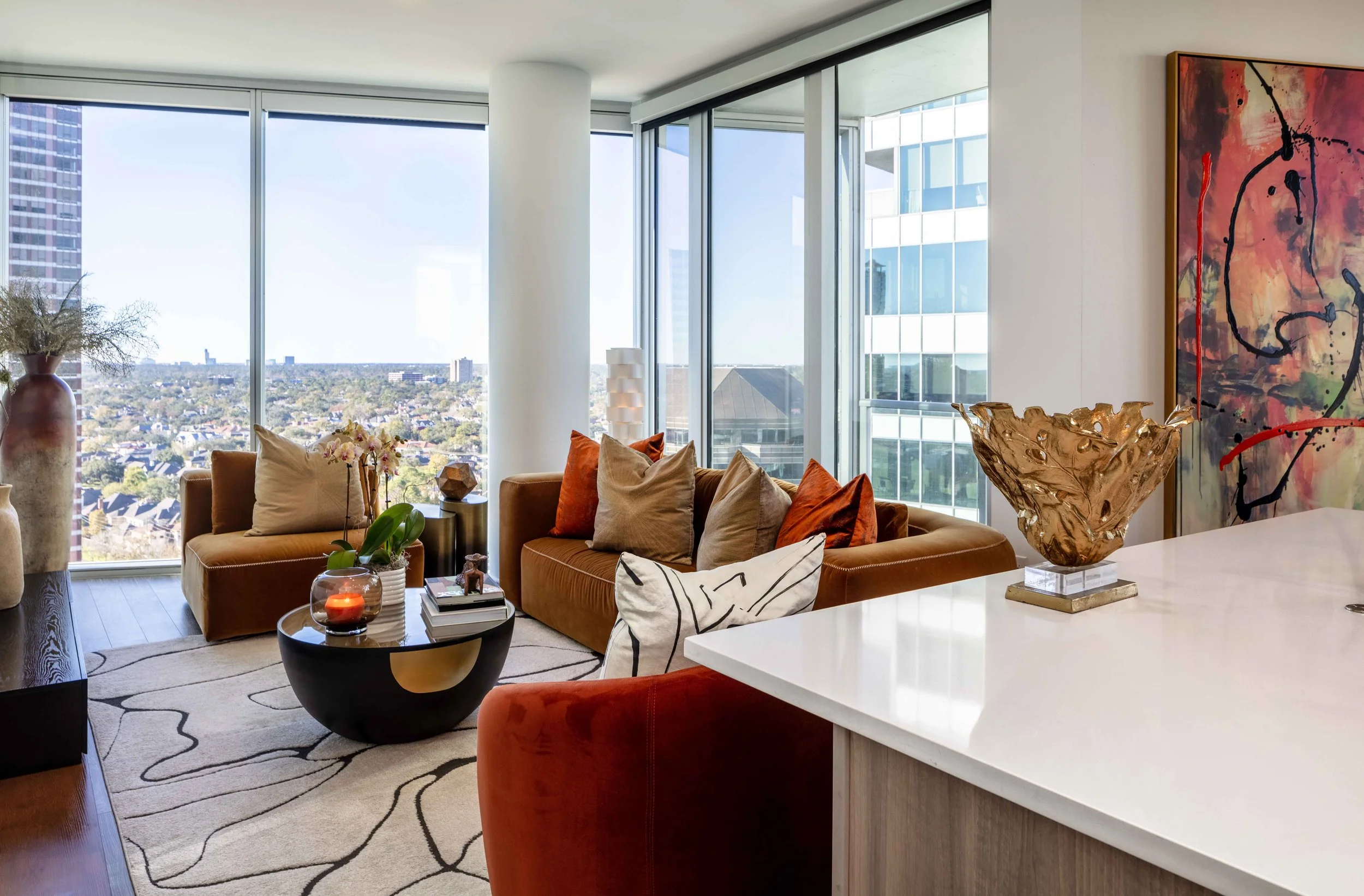 Open-concept living area with rust and camel velvet seating, modern decor, abstract artwork, and sweeping city views from large glass windows.