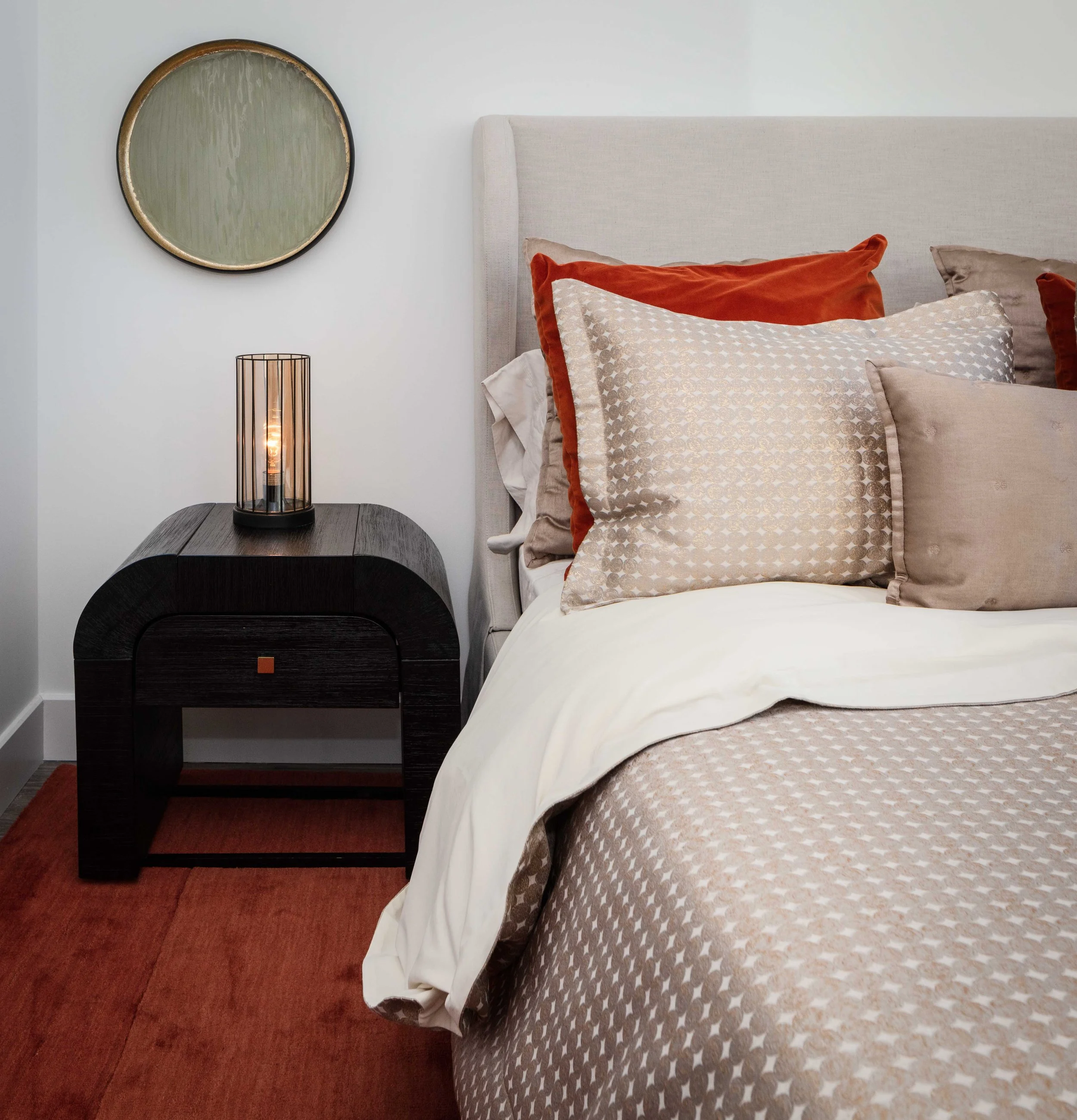Close-up of a neutral-toned bedroom with patterned bedding, rust accents, a round mirror, and a black nightstand with a modern lamp.