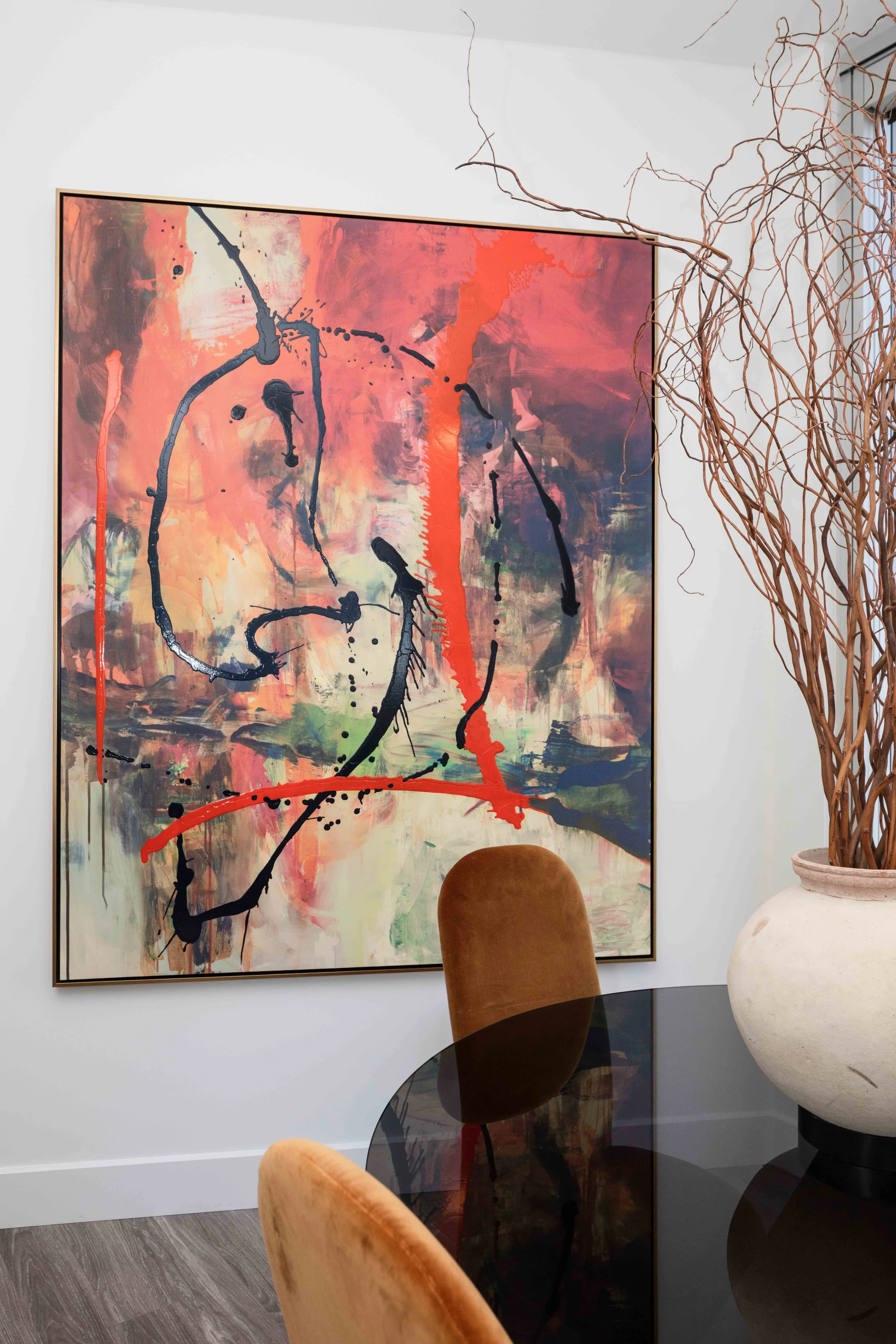 Vibrant abstract painting in orange and black tones above a modern dining table with rust velvet chairs and organic floral accents.