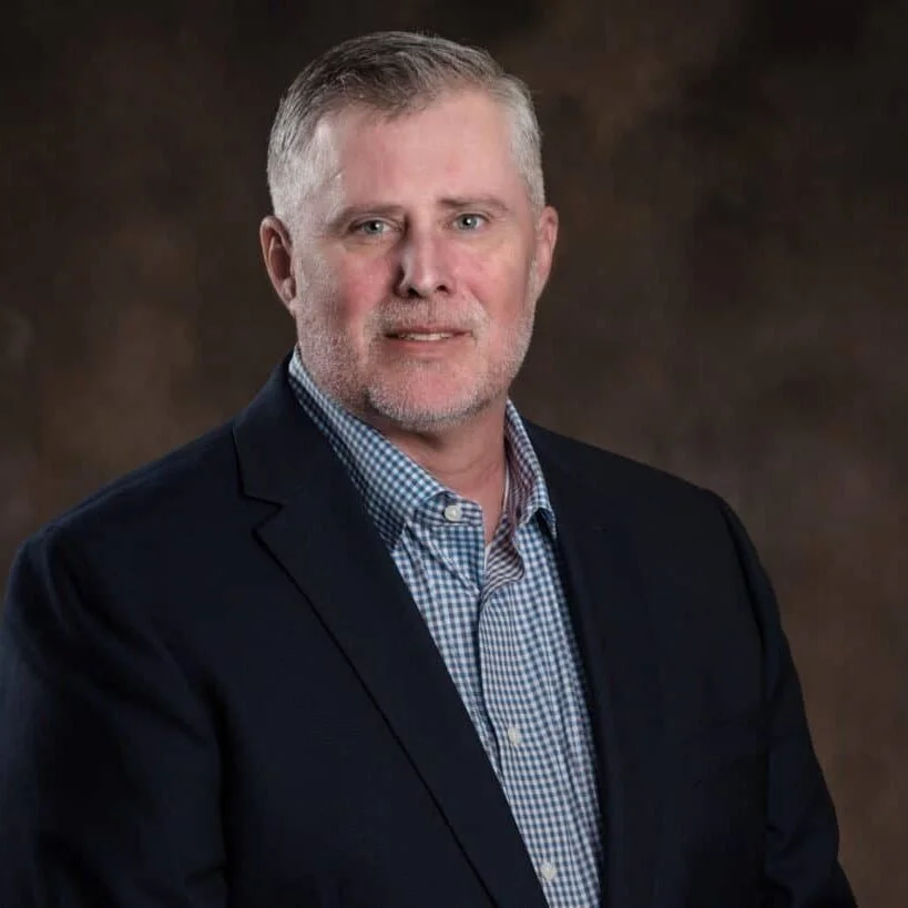 Middle-aged man wearing a dark suit and checkered shirt, posing in front of a dark, blurred background.