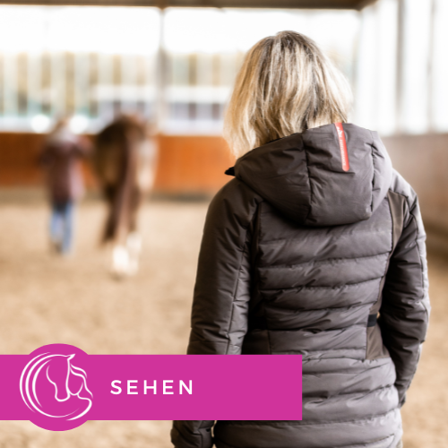 Eine Frau mit blondem Haar, die eine braune Winterjacke trägt, steht in einer Reithalle. Im Hintergrund ist ein Pferd sichtbar. Sie beobachtet die Bewegungsabläufe des Pferdes.