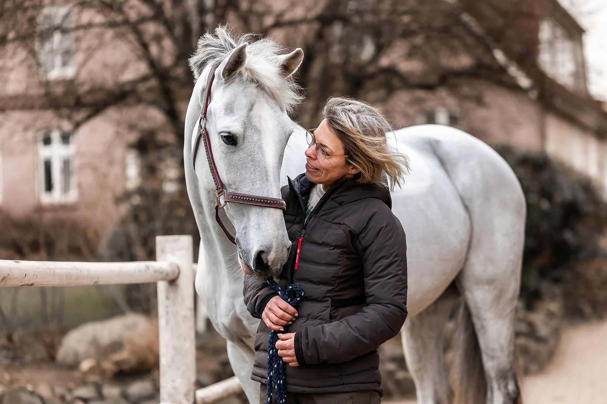 Eine Frau hält einen Schimmel am Halfter.  Es ist Herbst; die Bäume im Hintergrund sind kahl.