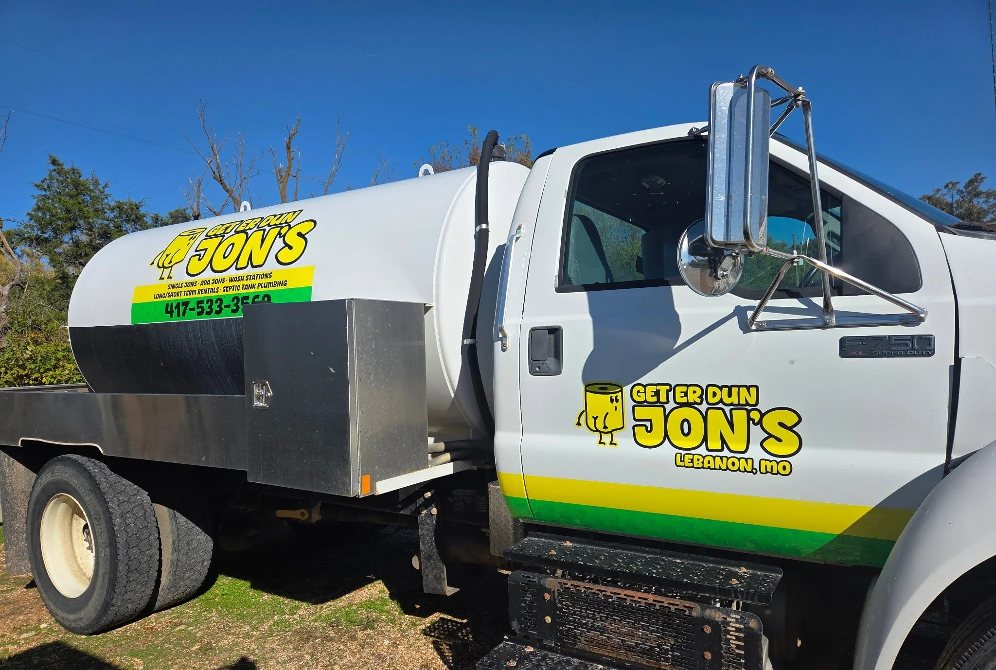 A white service truck with decals for Get Er Dun Jon's, a plumbing company in Lebanon, Missouri. The truck has a large tank and equipment for plumbing work, with the company's name, contact phone number, and services listed.