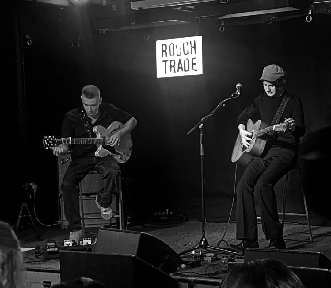 Two musicians playing acoustic guitars on a small stage with a sign that reads 'Rough Trade' in the background, in a black and white photo.